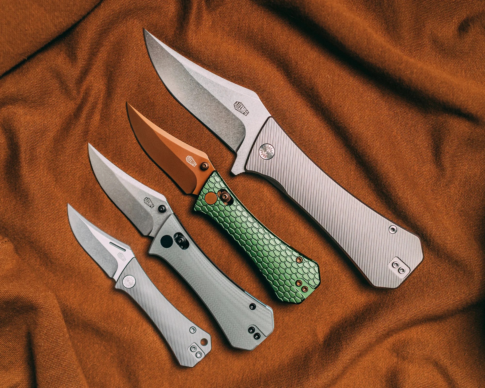 Five folding knives with metal handles and various blade finishes laid out on a brown fabric background.