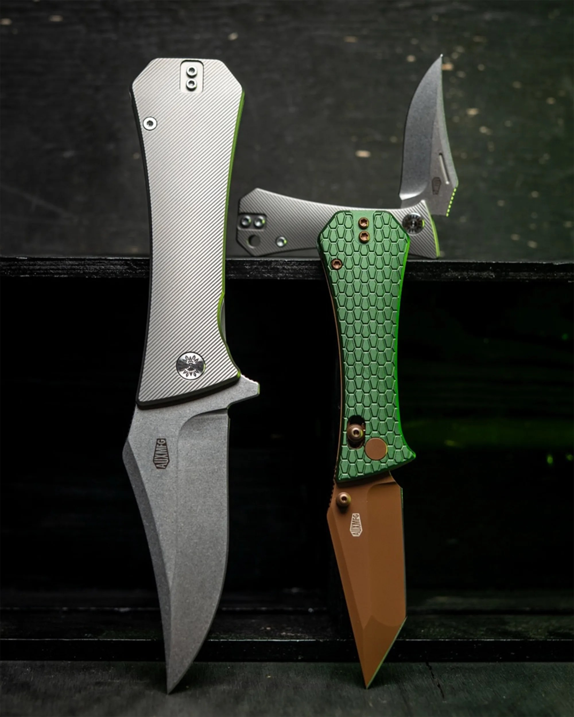 Two folding knives with textured handles, one silver with a stonewashed blade and one green with a brown blade, displayed on a black surface.