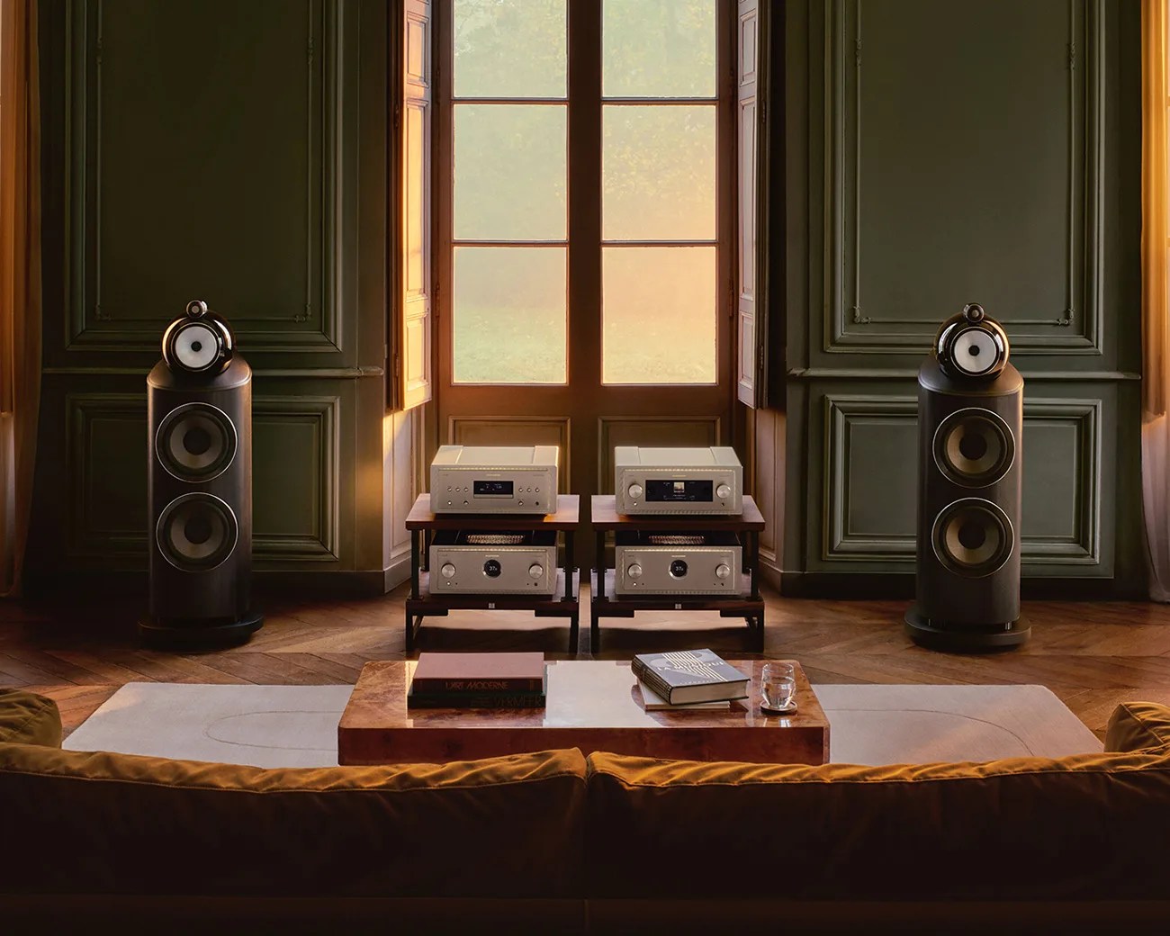 Pair of large black floor-standing speakers with white drivers flanking two wooden racks holding silver audio components in a room with green paneled walls.