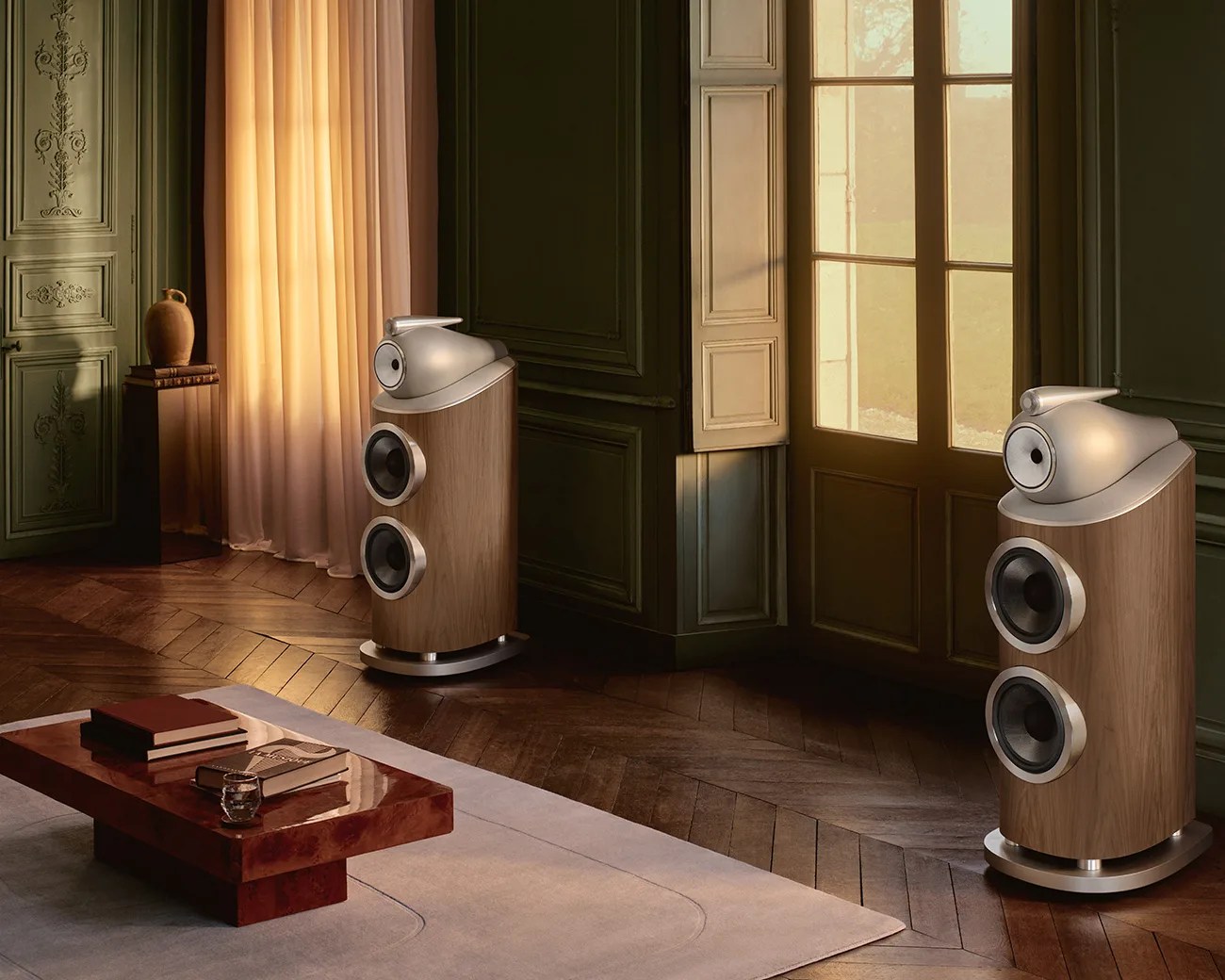 Pair of modern floor-standing speakers with wood finish and silver top sections in a warmly lit room with wooden floors and a low wooden table.