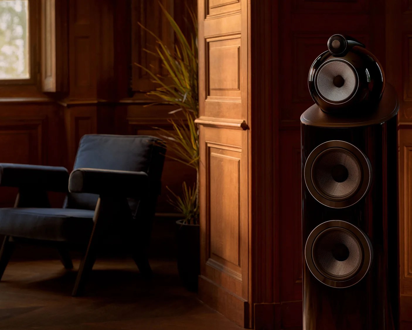 Glossy black floor-standing speaker with three drivers in a wood-paneled room next to a dark upholstered armchair.