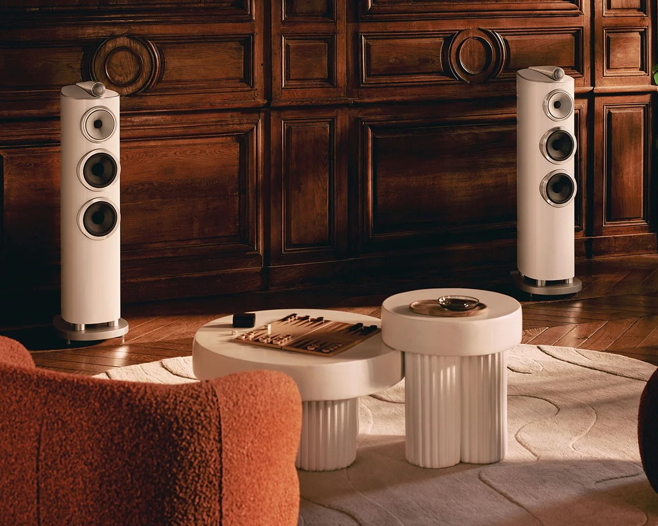 Pair of tall, beige floor-standing speakers with three drivers each in a room with dark wood paneling and modern white coffee tables.