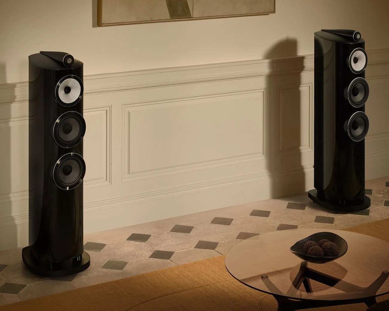 Pair of tall black floor-standing speakers with white and black drivers in a beige room with patterned tile floor.