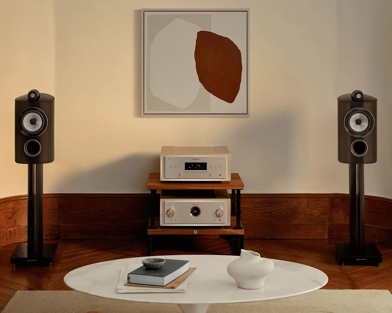 Two black Bowers & Wilkins speakers on stands flanking a wooden shelf with beige Marantz audio components below abstract wall art.