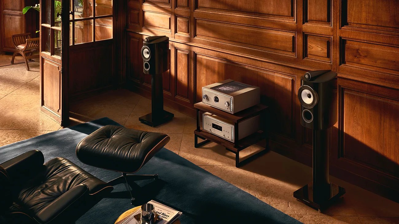 Black floor-standing speakers with white drivers and silver audio components on a wooden stand against a wood-paneled wall.