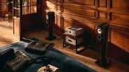 Black floor-standing speakers with white drivers and silver audio components on a wooden stand against a wood-paneled wall.
