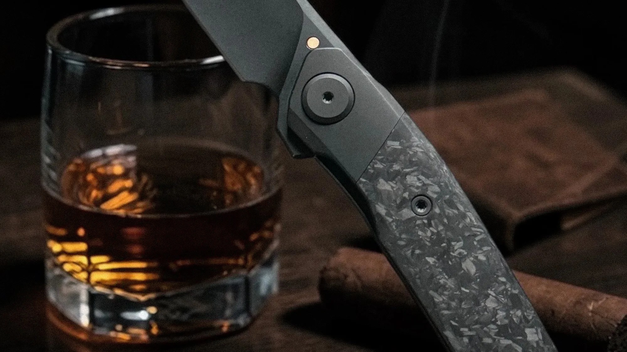 Close-up of a textured gray folding knife handle next to a glass of amber liquid on a wooden surface.