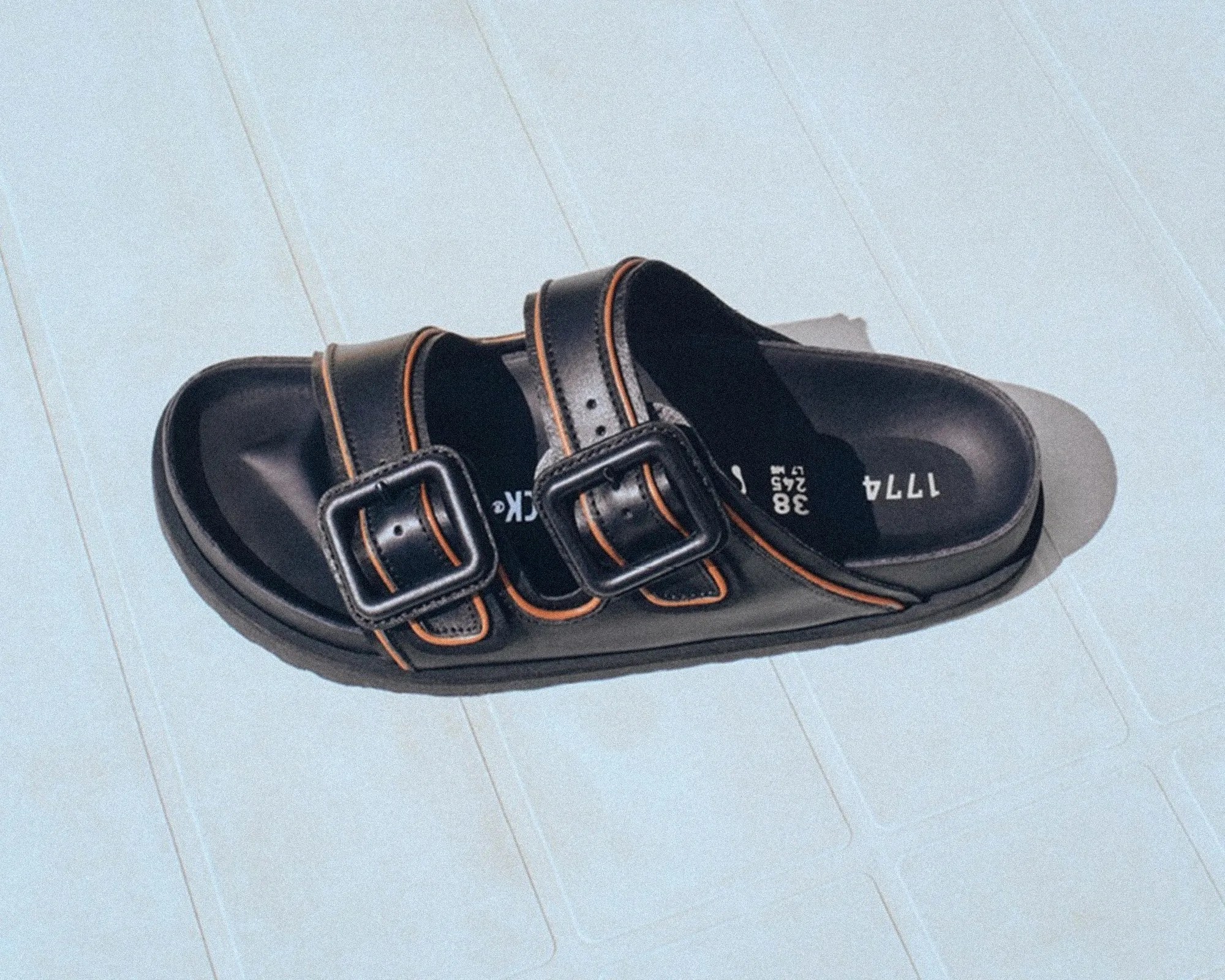 Pair of black leather sandals with double buckles and orange stitching on a light blue surface.