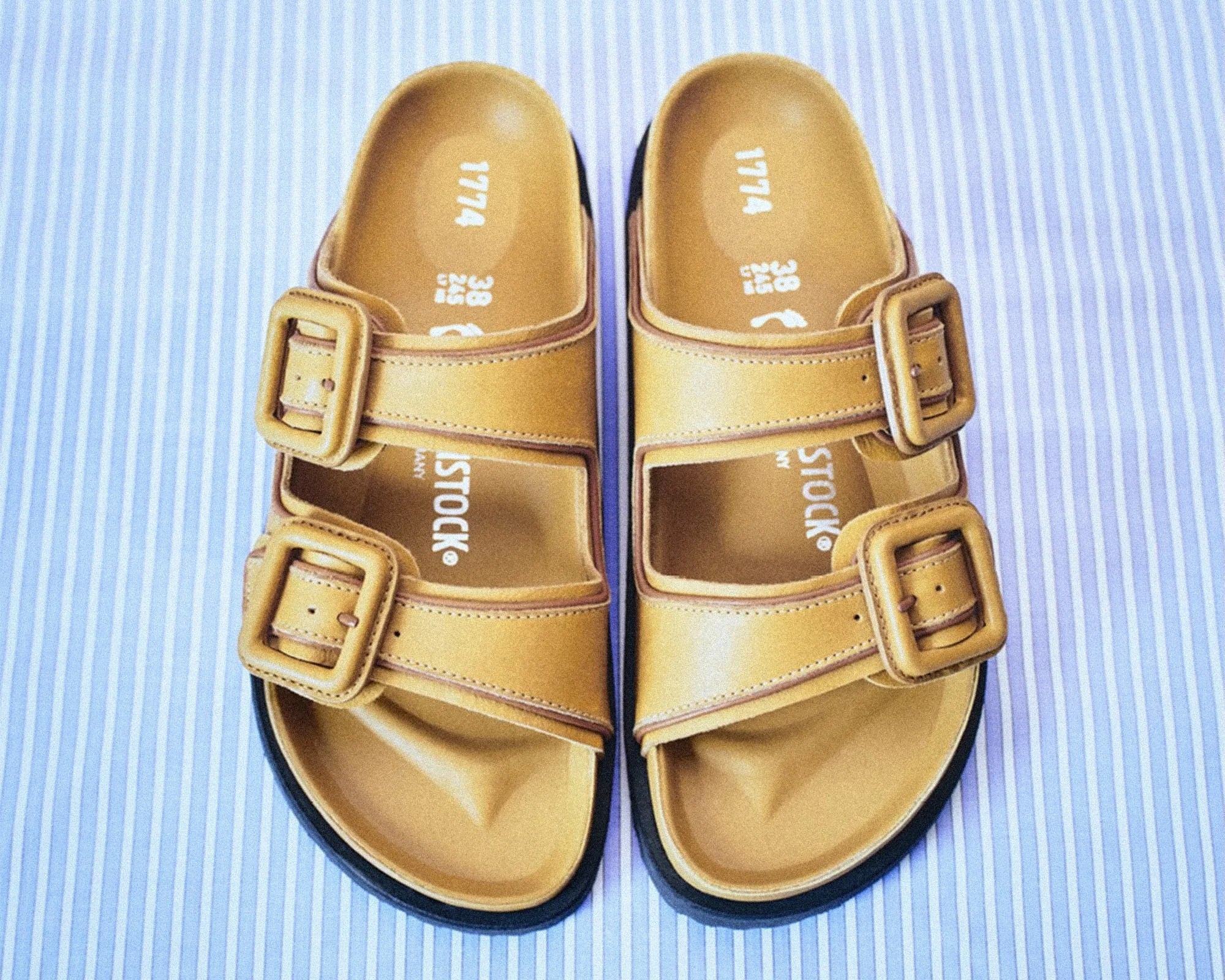Pair of tan leather sandals with double buckle straps on a blue and white striped fabric background.