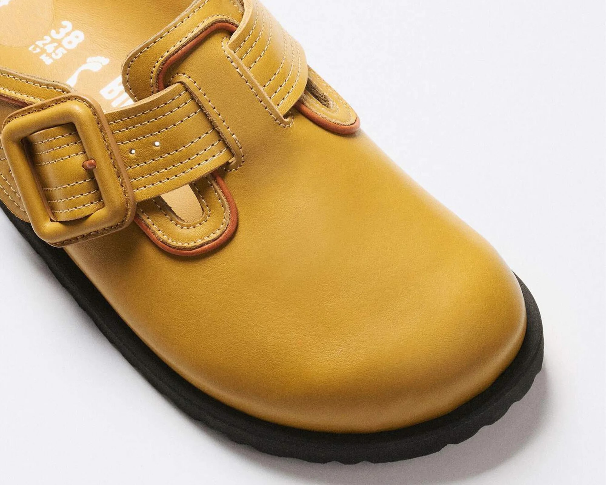 Close-up of a mustard yellow leather shoe with a matching buckle and black sole.