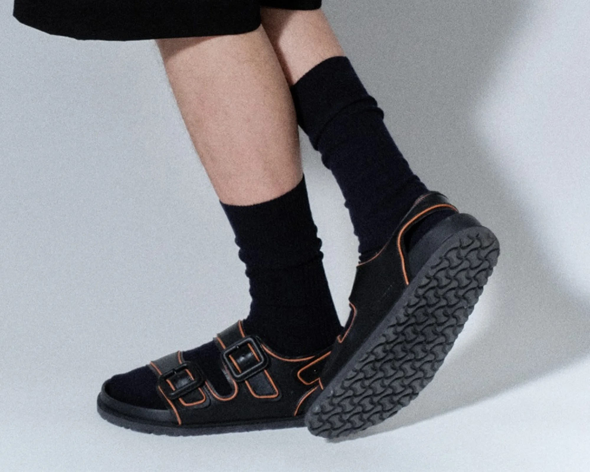 Black sandals with orange trim and three buckles worn with black socks on crossed legs.