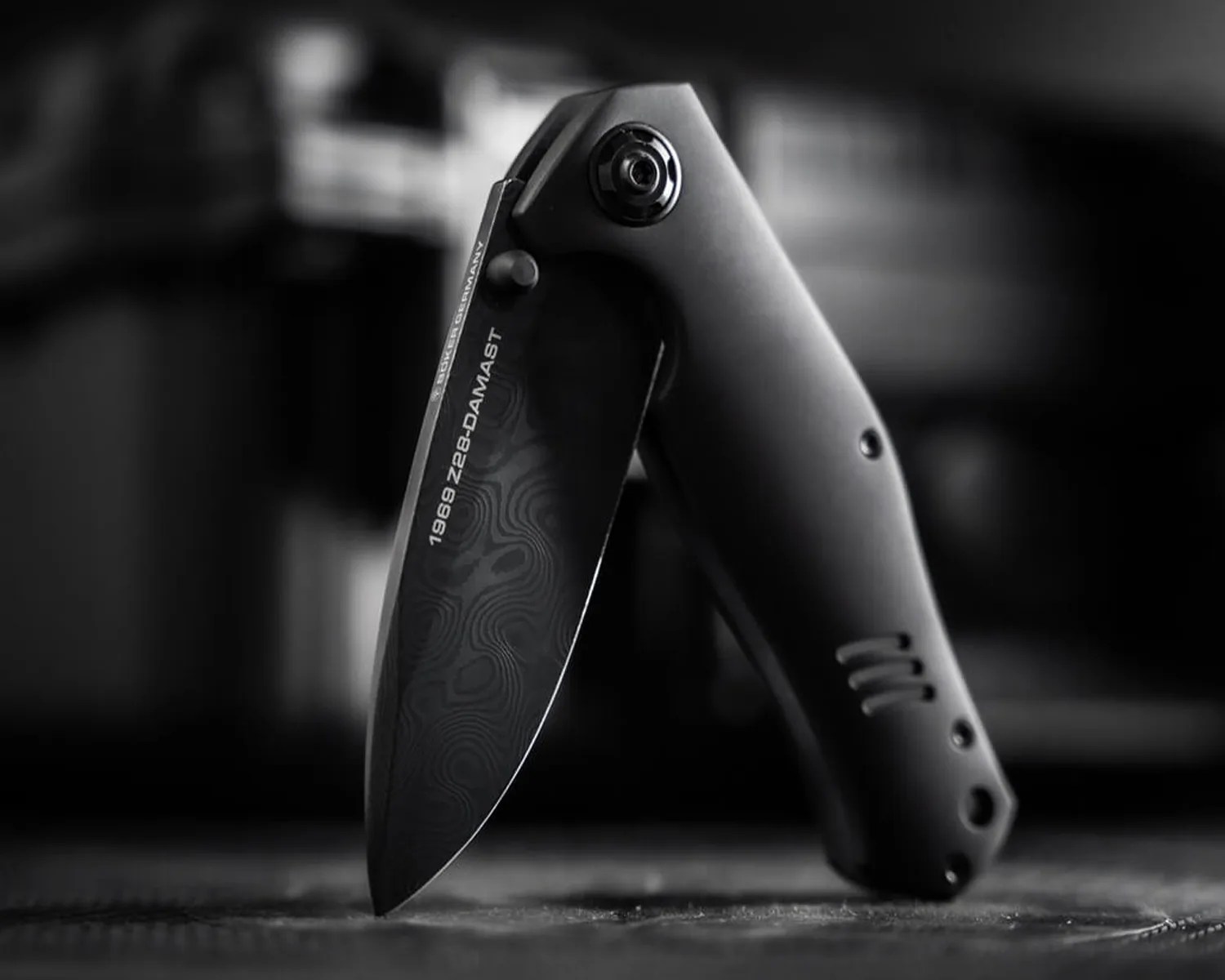 Black folding knife with patterned blade and "1969 Z28-DAMAST" text on the blade, standing on a textured surface.
