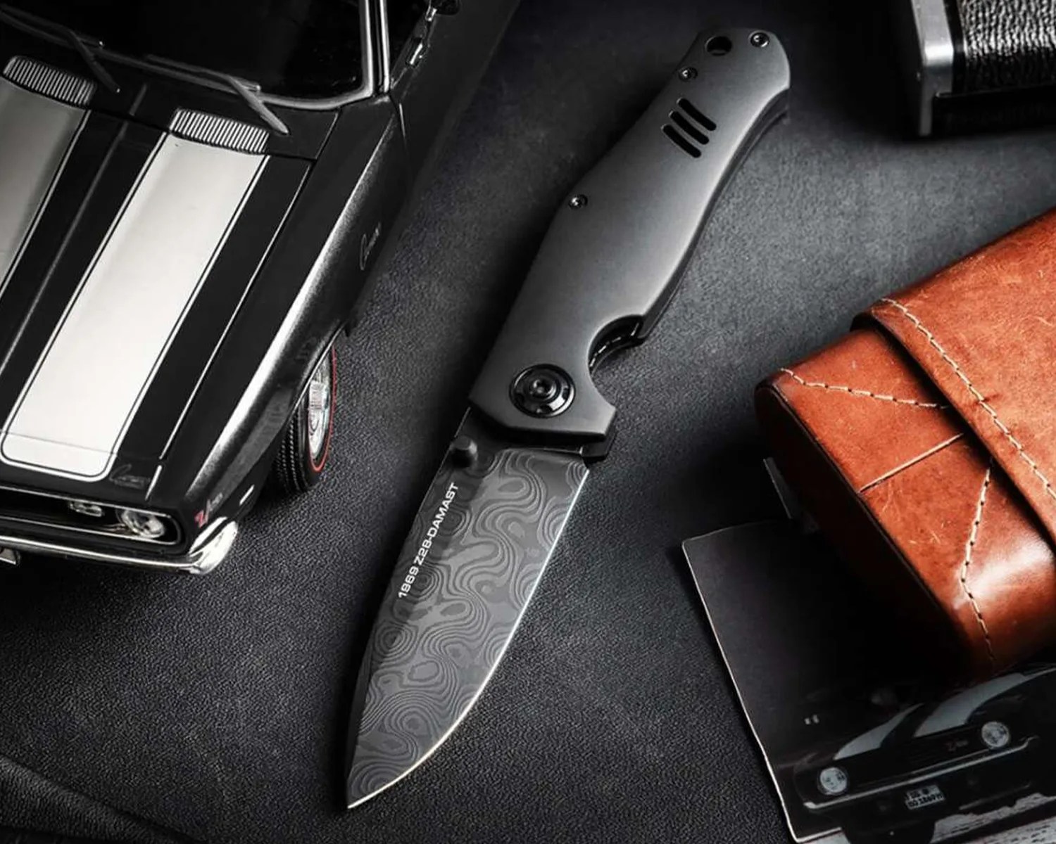 Black folding knife with patterned blade and matte handle on a dark surface next to a brown leather wallet and a black model car.