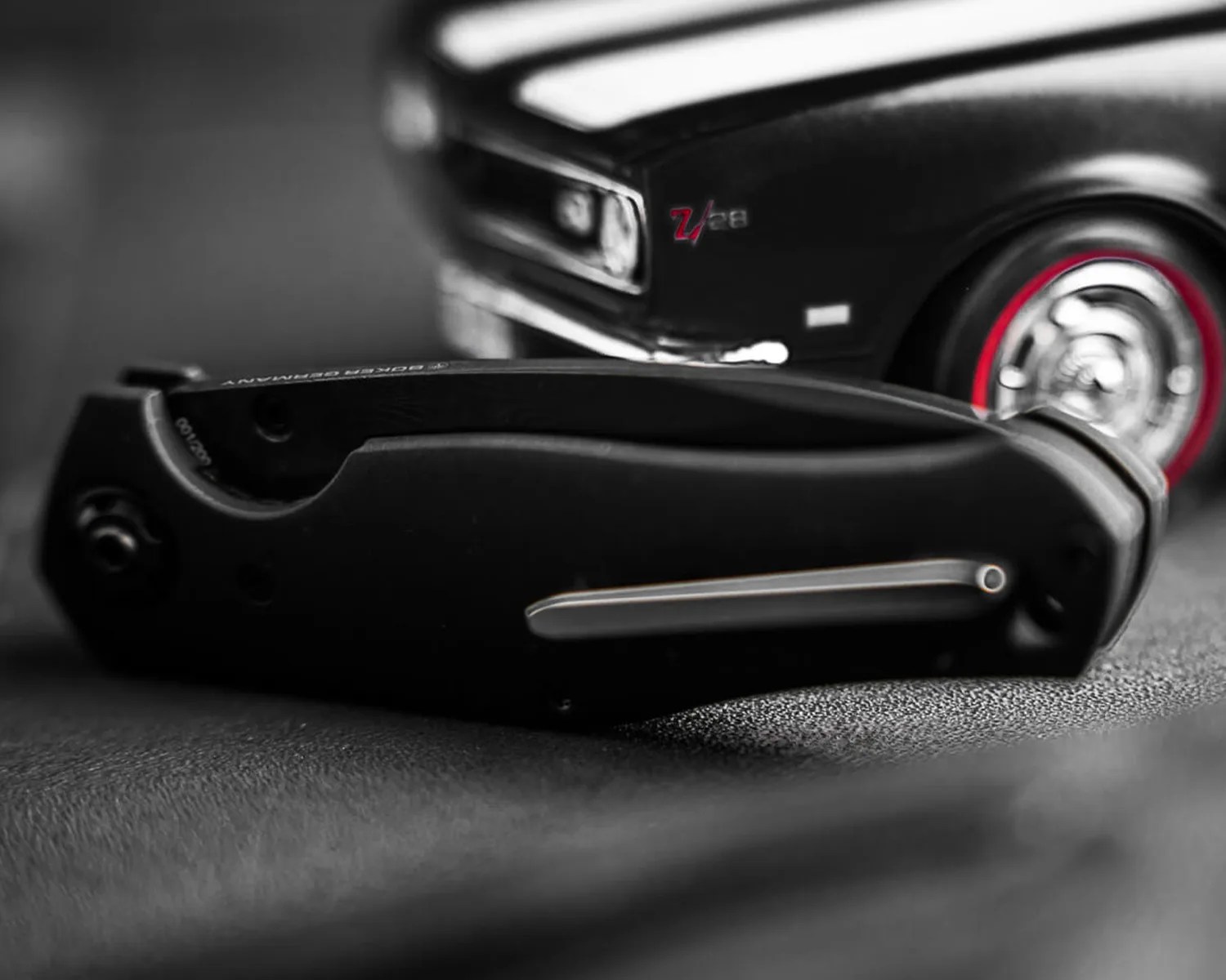 Black folding pocket knife with a silver clip lying on a textured surface, with a blurred black car model in the background.