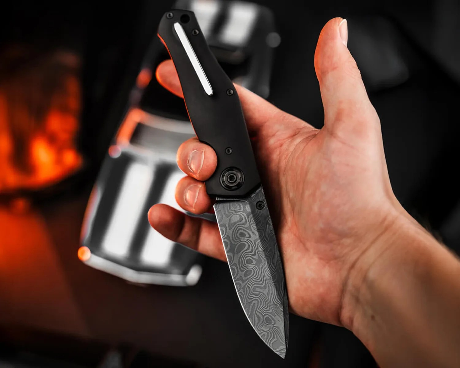 Hand holding a black folding knife with a patterned Damascus steel blade and a silver clip.