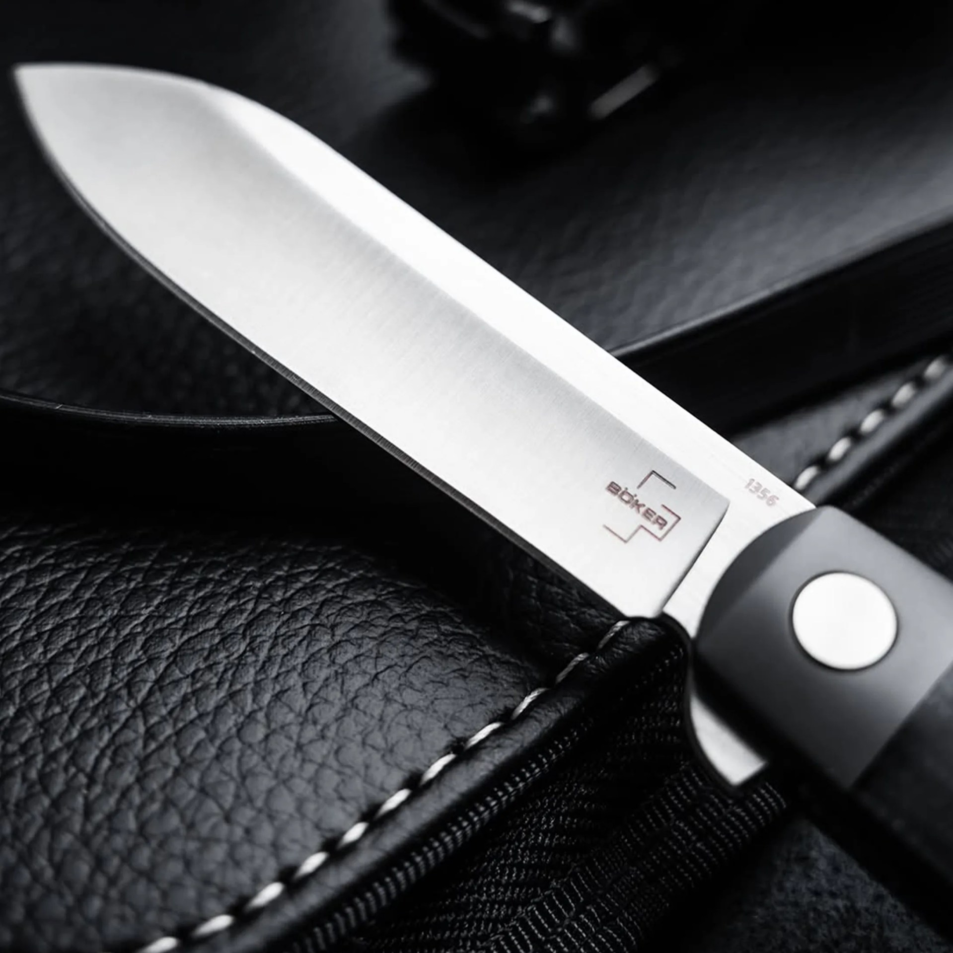 Close-up of a Böker knife blade with a black handle resting on textured black leather.