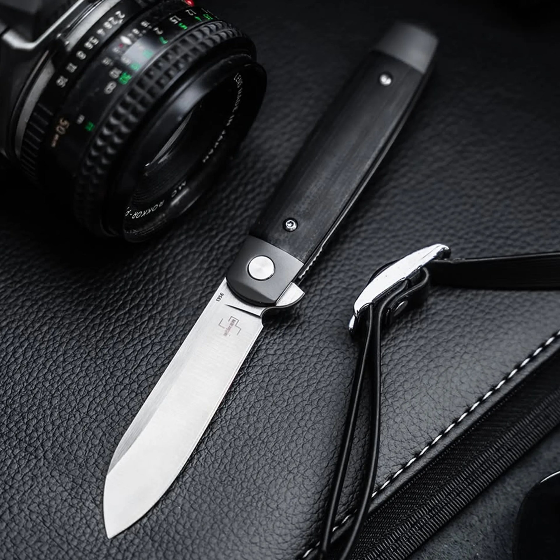 Black-handled folding knife with a partially visible camera lens and black leather surface background.