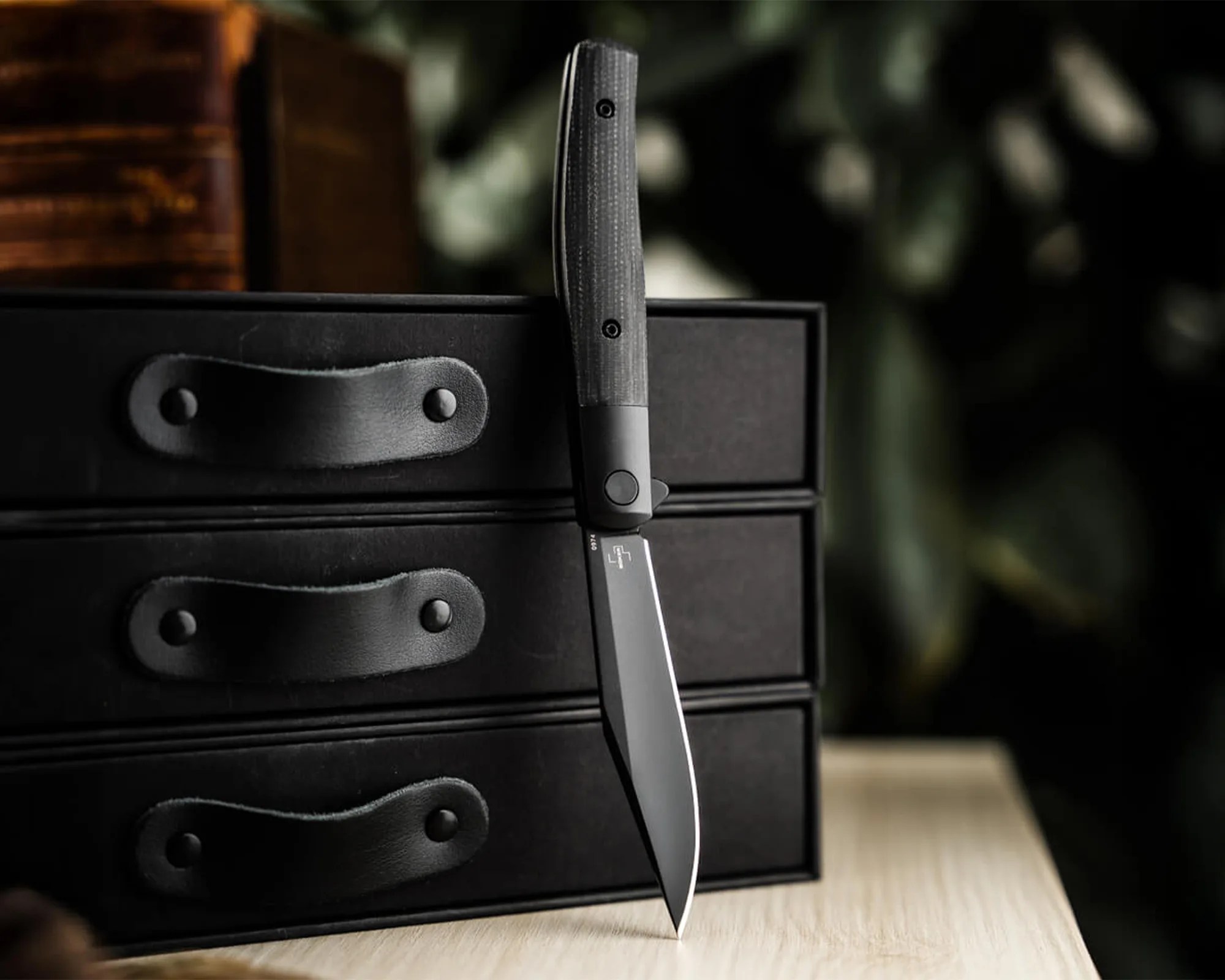 Black knife with textured handle standing vertically against a black box with leather handles on a wooden surface.