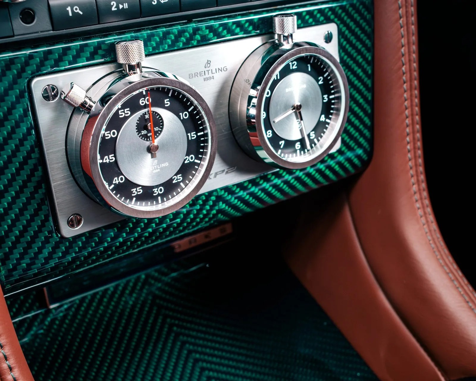 Close-up of two chrome Breitling stopwatch dials mounted on a green carbon fiber panel with brown leather trim.