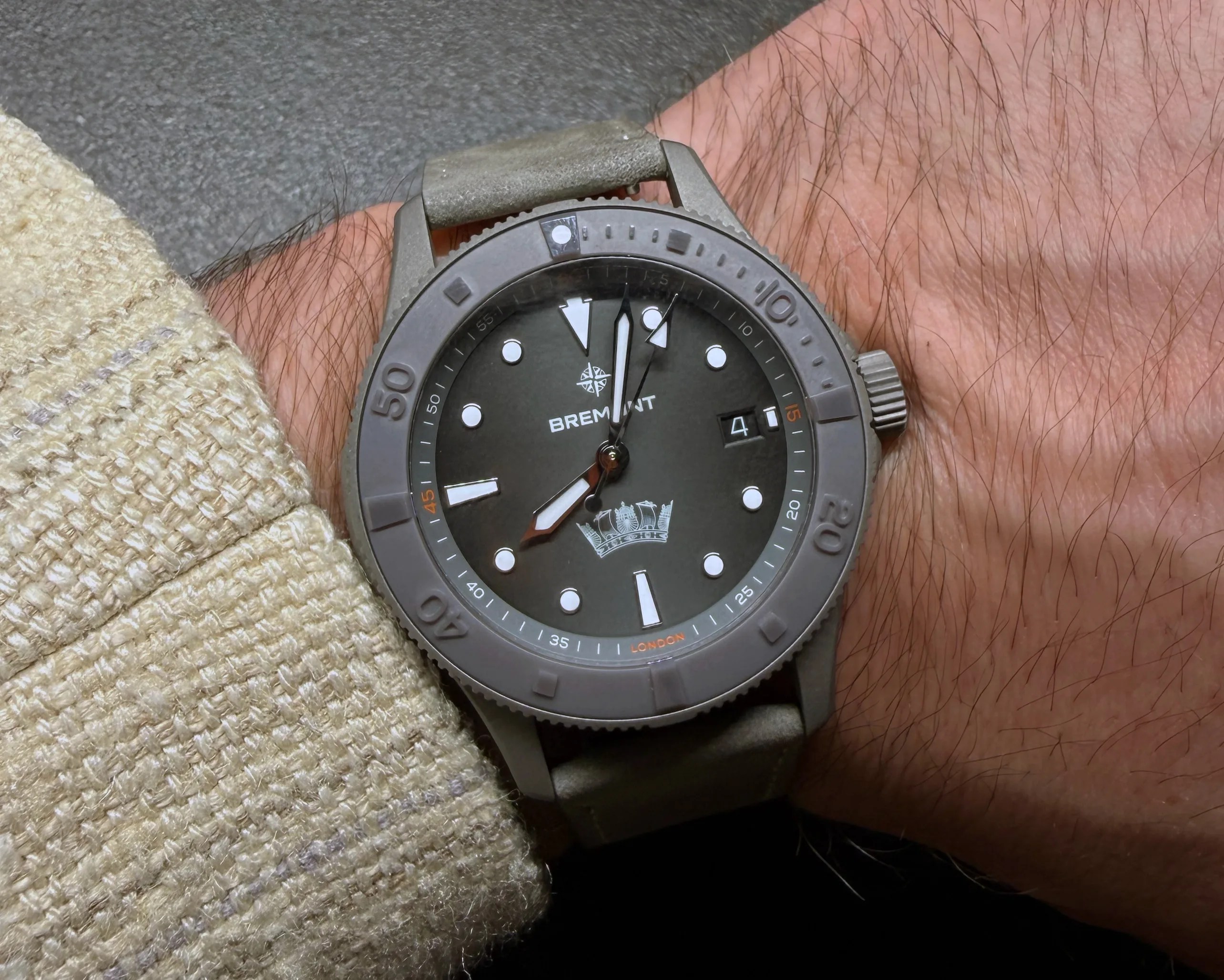 Gray Bremont wristwatch with round dial and white markers on a hairy wrist wearing a beige textured sleeve.