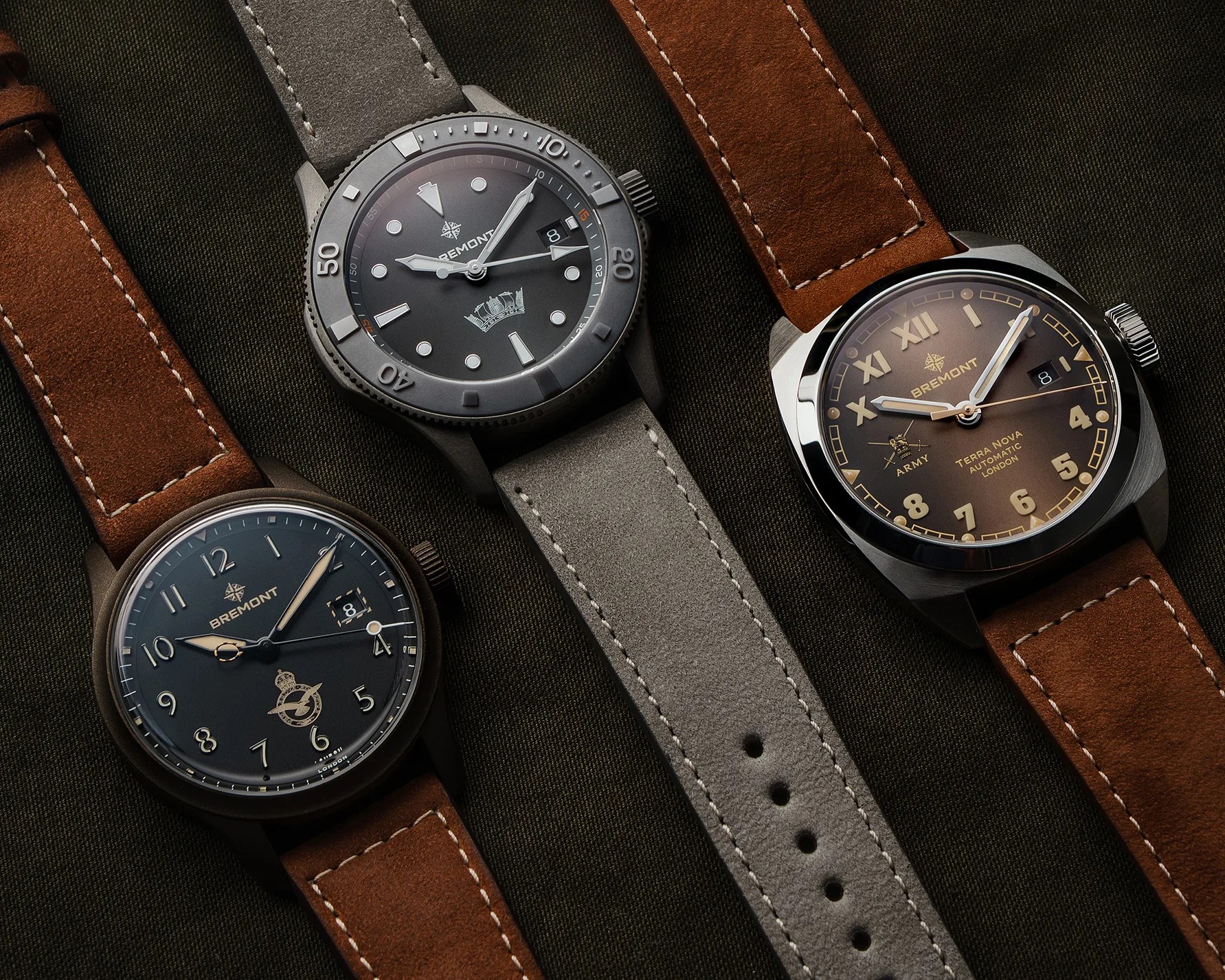 Three Bremont wristwatches with brown and gray leather straps on a dark fabric background.