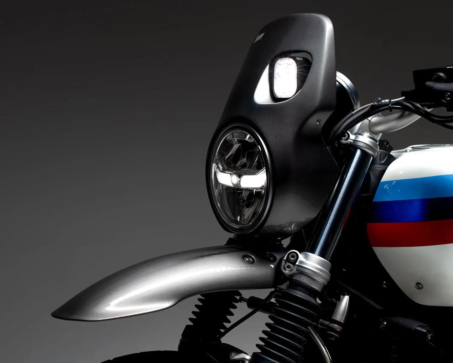 Close-up of a motorcycle front with a round LED headlight, black fairing, silver front fender, and a fuel tank with red, blue, and light blue stripes.