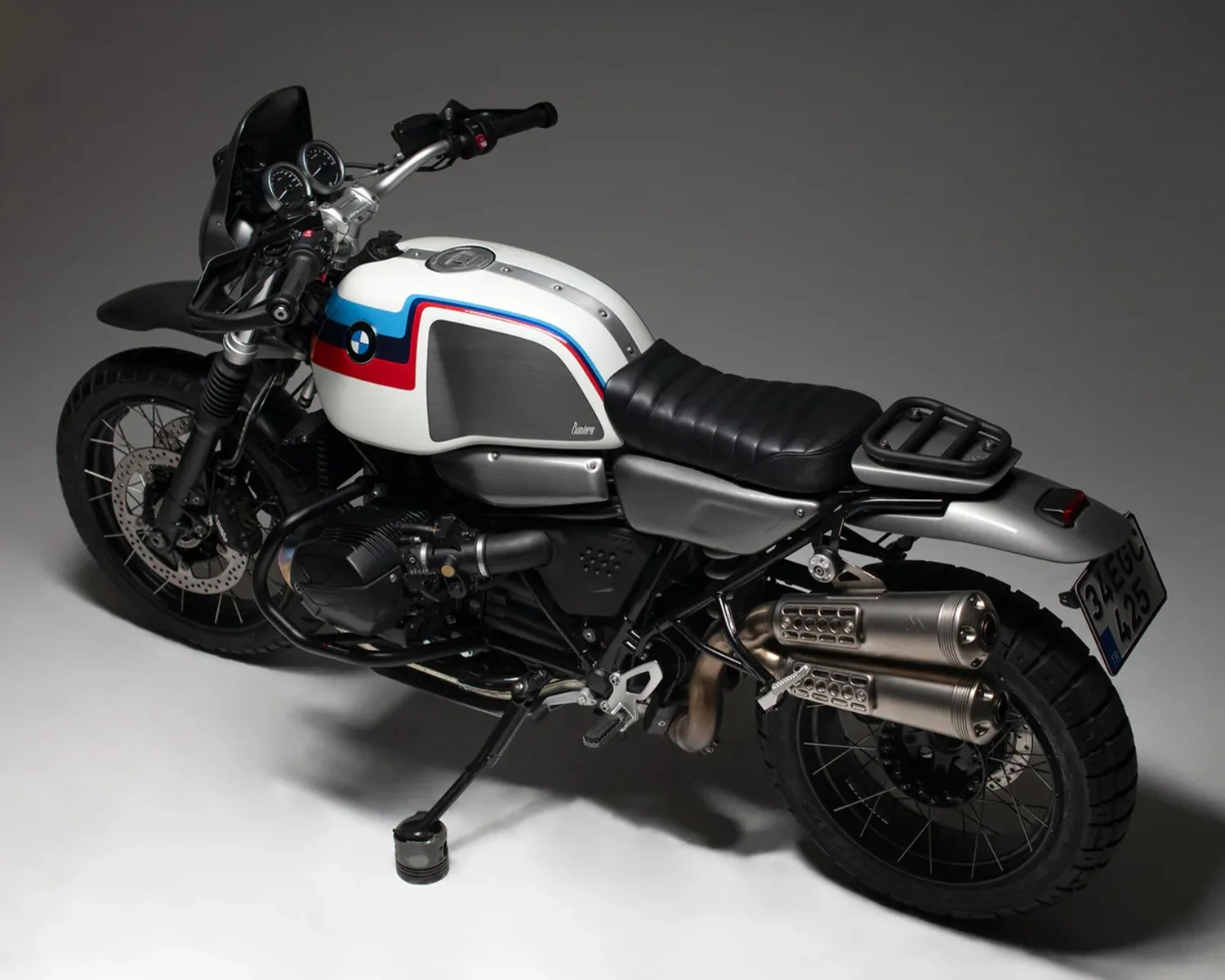 White BMW motorcycle with red and blue stripes, black seat, dual exhaust, and knobby tires on a gray background.