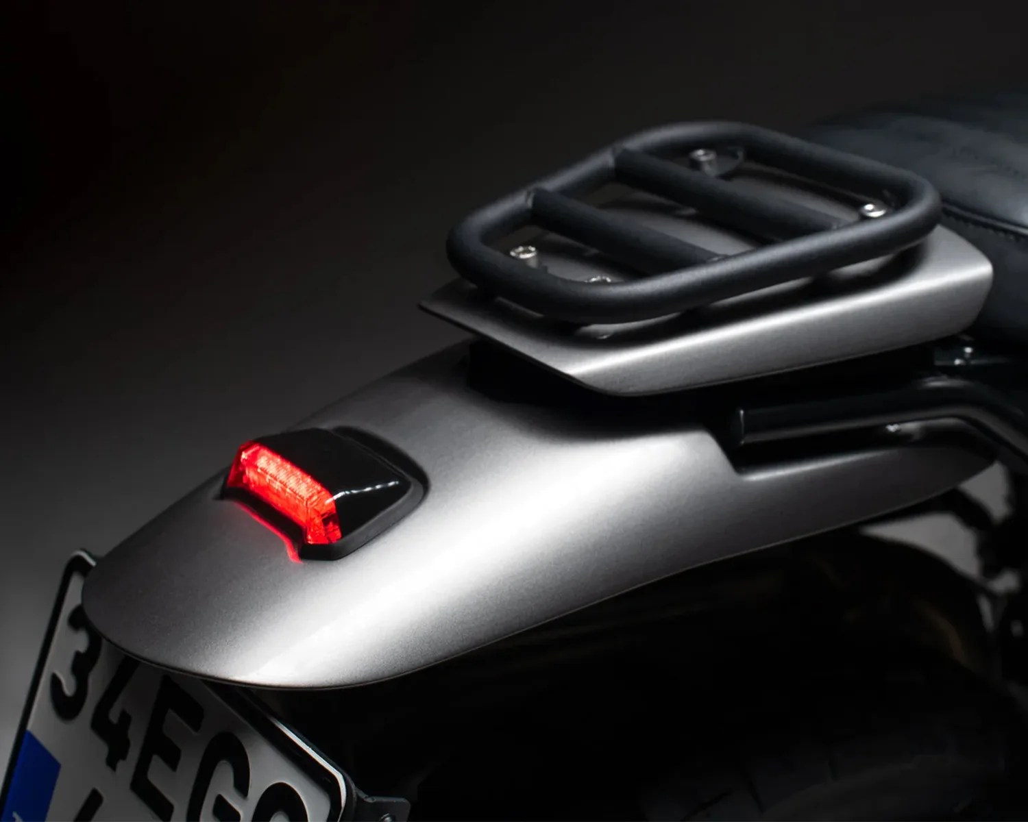 Rear section of a motorcycle with a matte silver fender, red tail light, black luggage rack, and partially visible license plate.