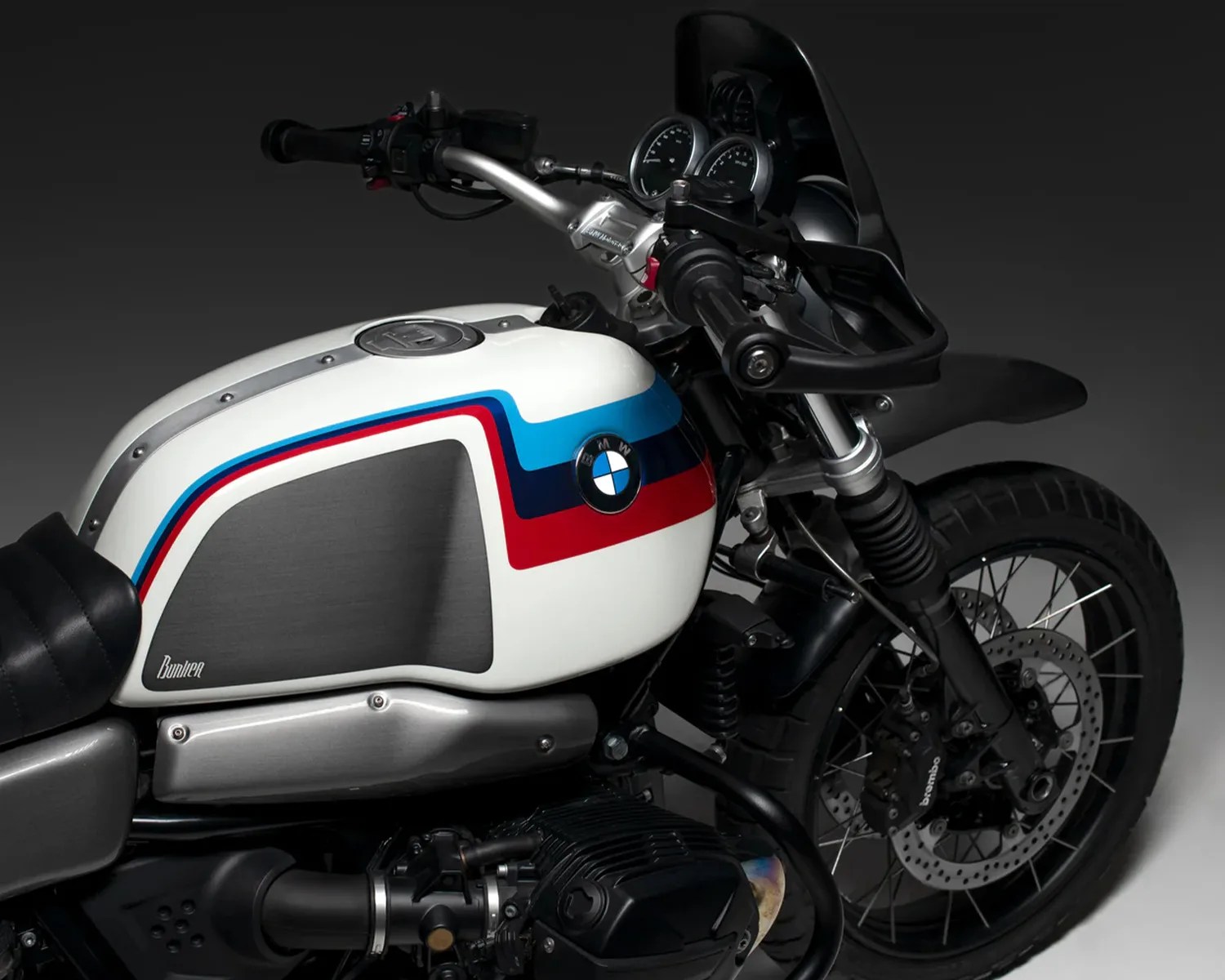 Close-up of a white BMW motorcycle fuel tank with red, blue, and light blue stripes and black front wheel with Brembo brake caliper.