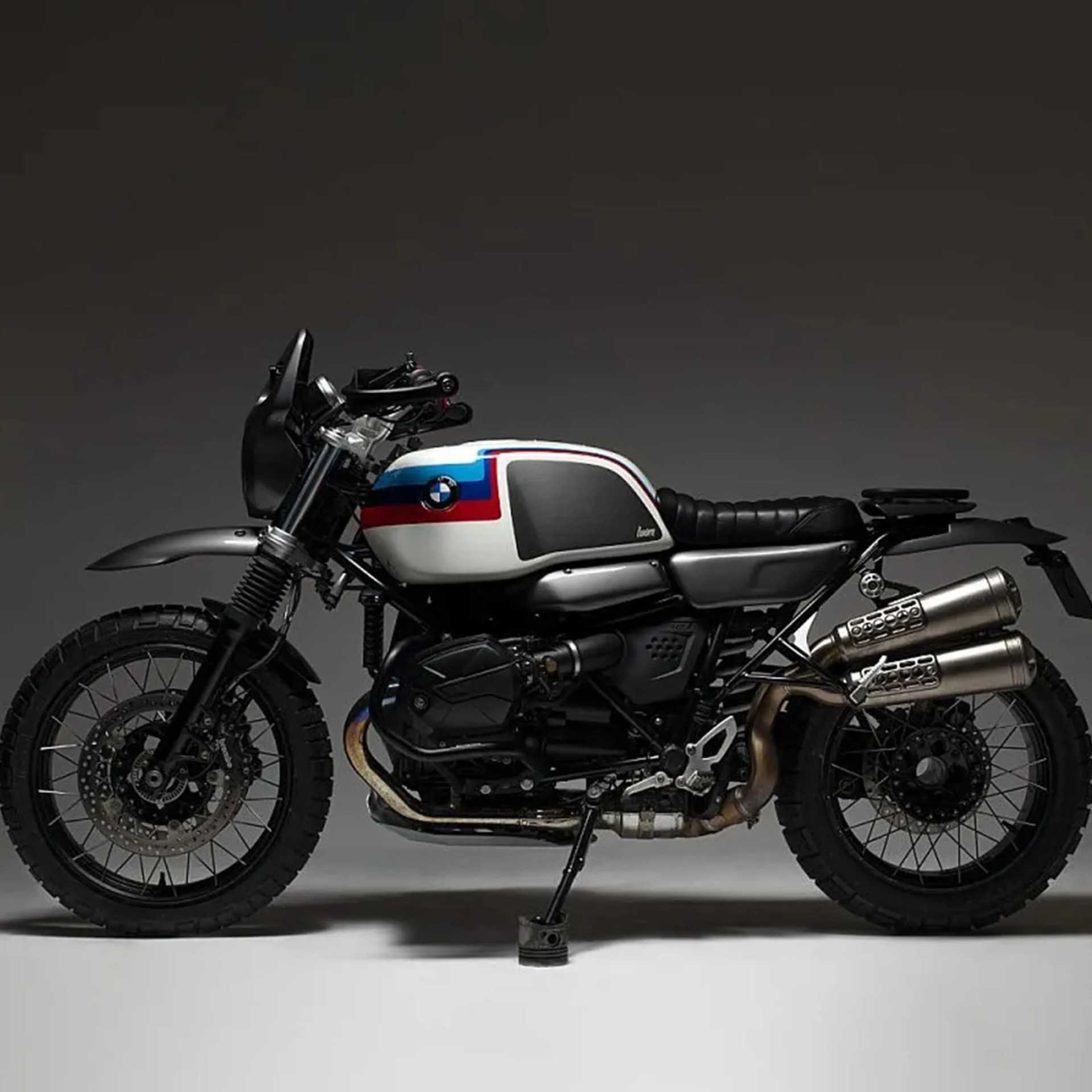 White BMW motorcycle with blue and red stripes, black seat, and dual exhaust pipes on a gray background.