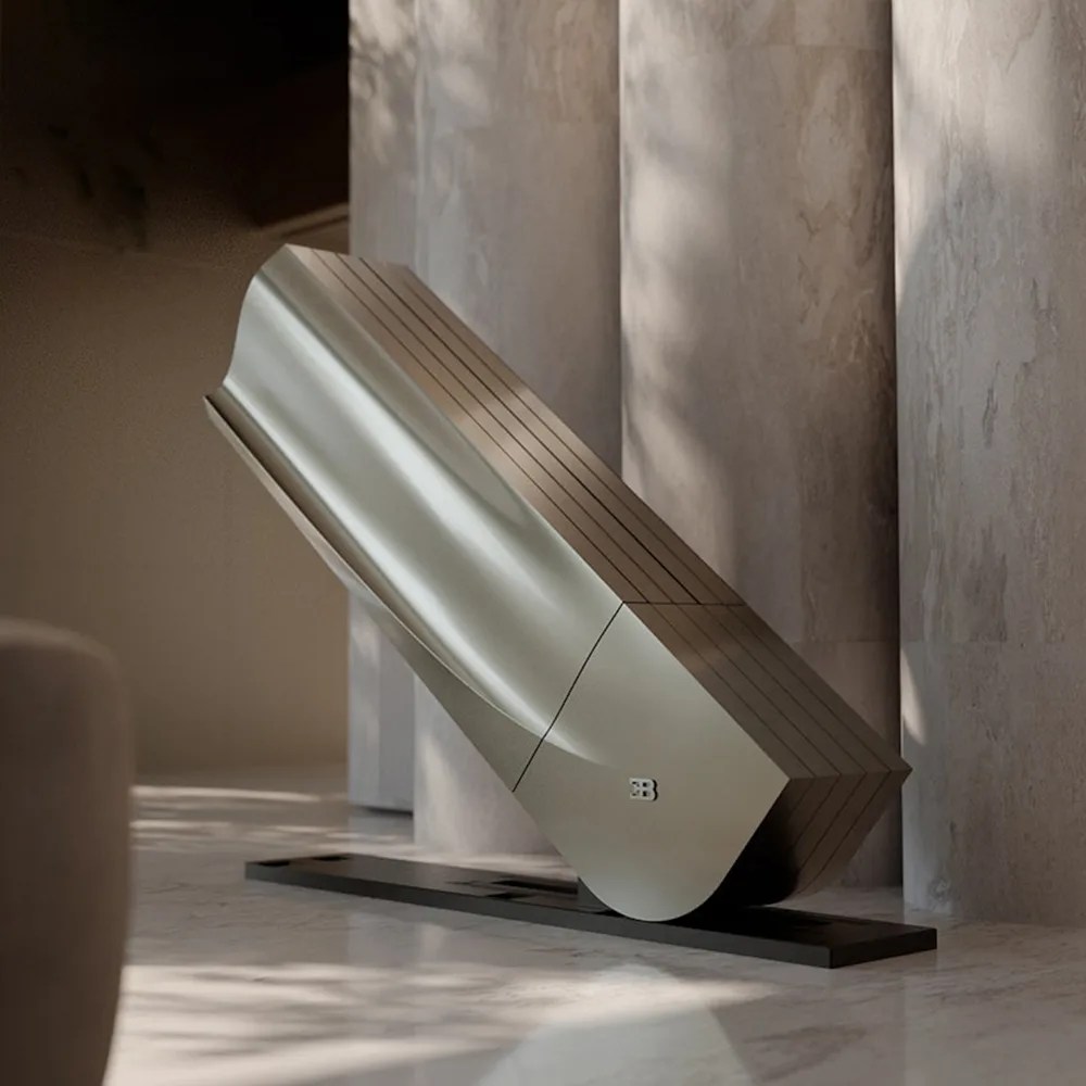 Sleek metallic sculpture with a smooth curved surface and horizontal lines, mounted on a black base against a stone wall.