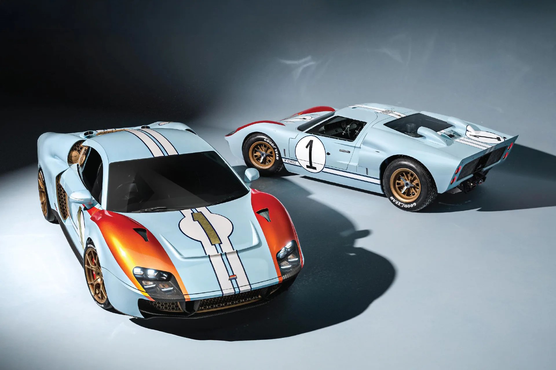 Two light blue race cars with white stripes and gold wheels, one with orange fenders and the other with red accents, on a gray floor.