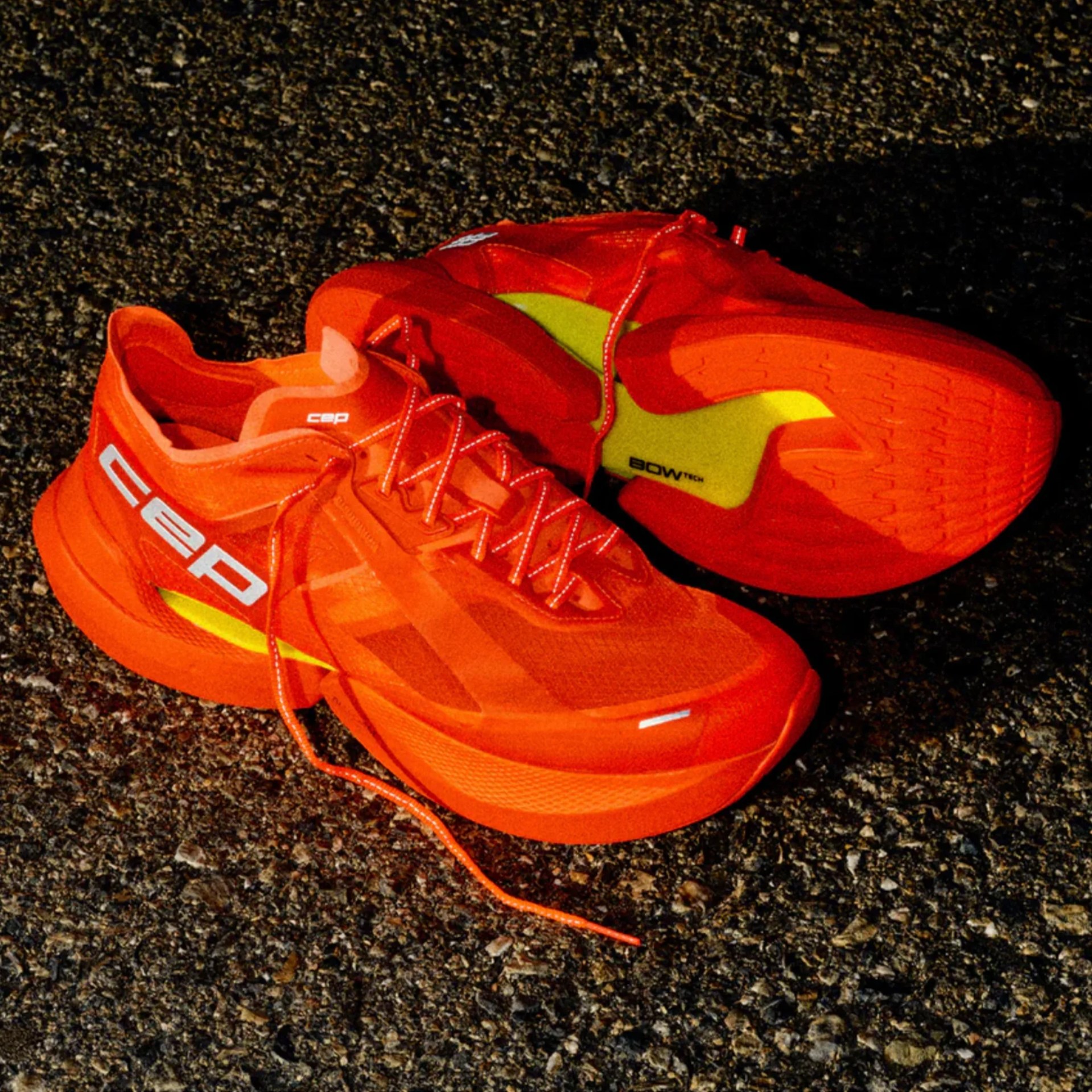 Bright orange running shoes with white "cep" branding and yellow "BOWTECH" detail on a textured asphalt surface.