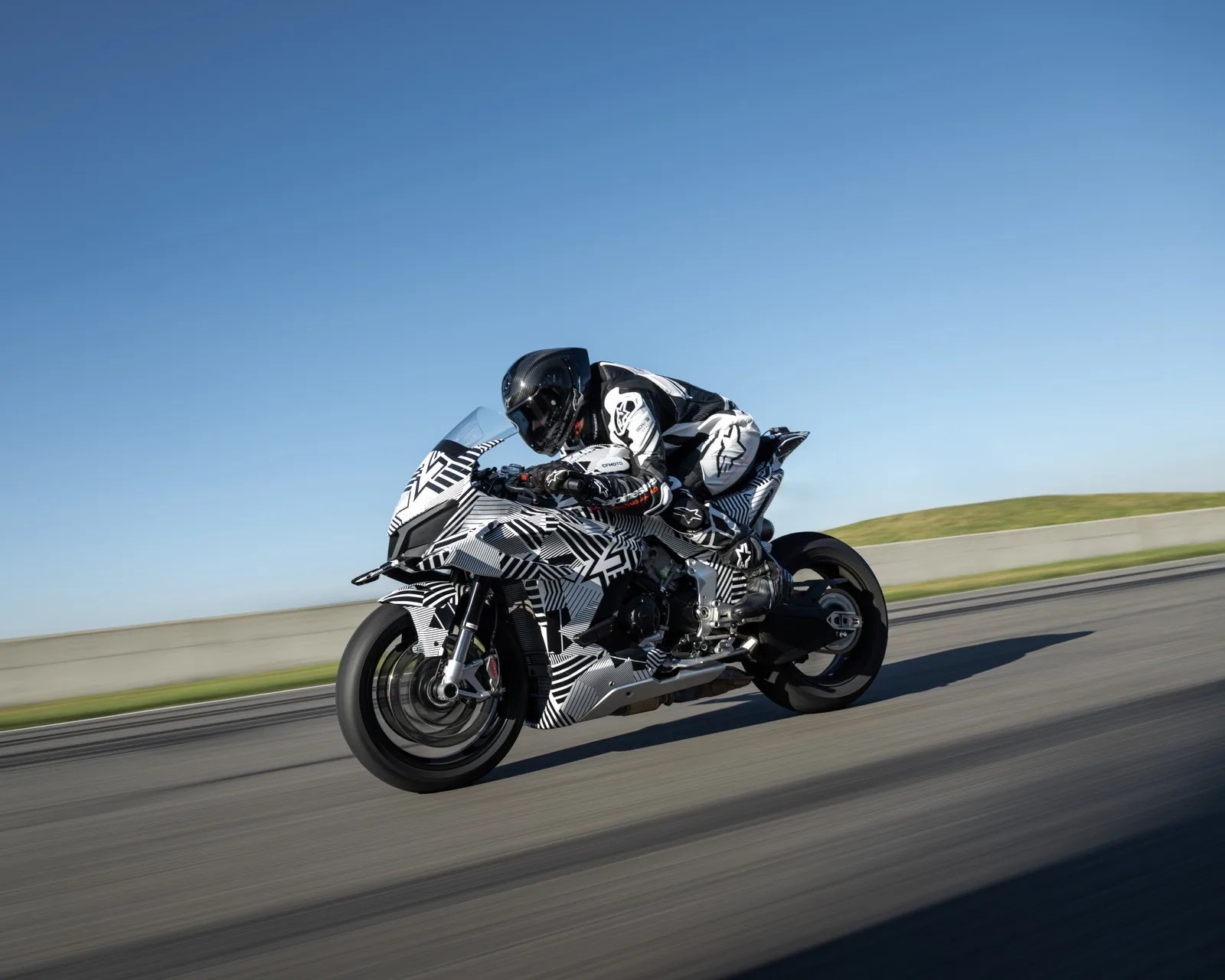 Motorcyclist in black and white gear riding a black and white patterned sportbike on a racetrack under clear blue sky.