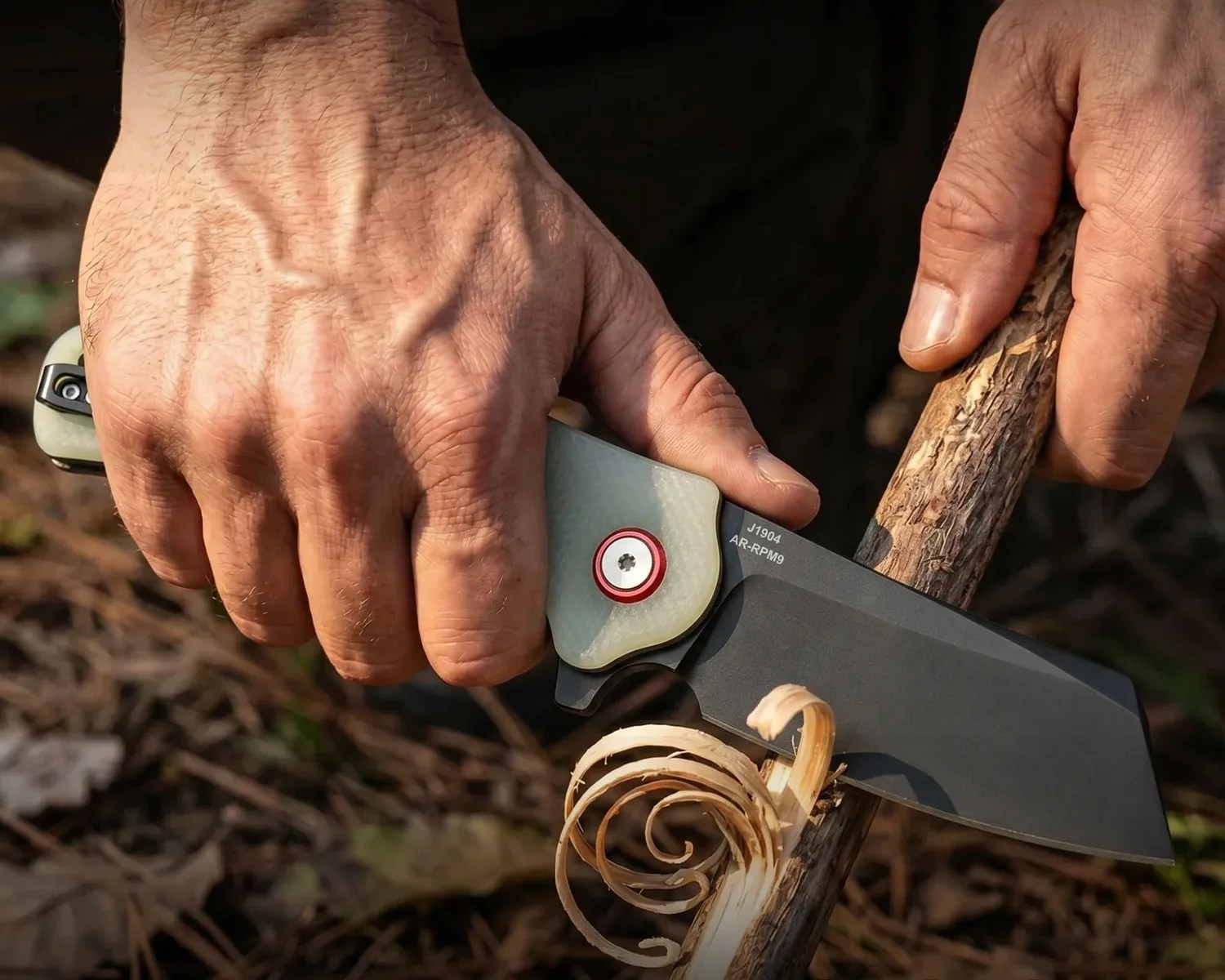Hands whittling a stick with a black folding knife marked J1904 AR-RPM9.