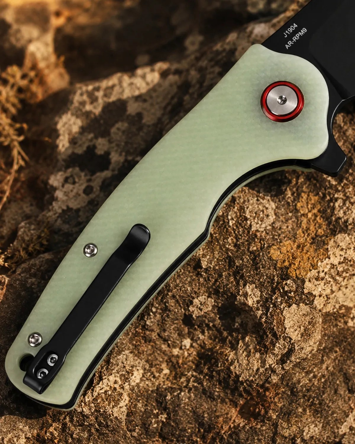 Folding knife with a pale green textured handle, black pocket clip, and red-accented pivot screw on a rocky surface.