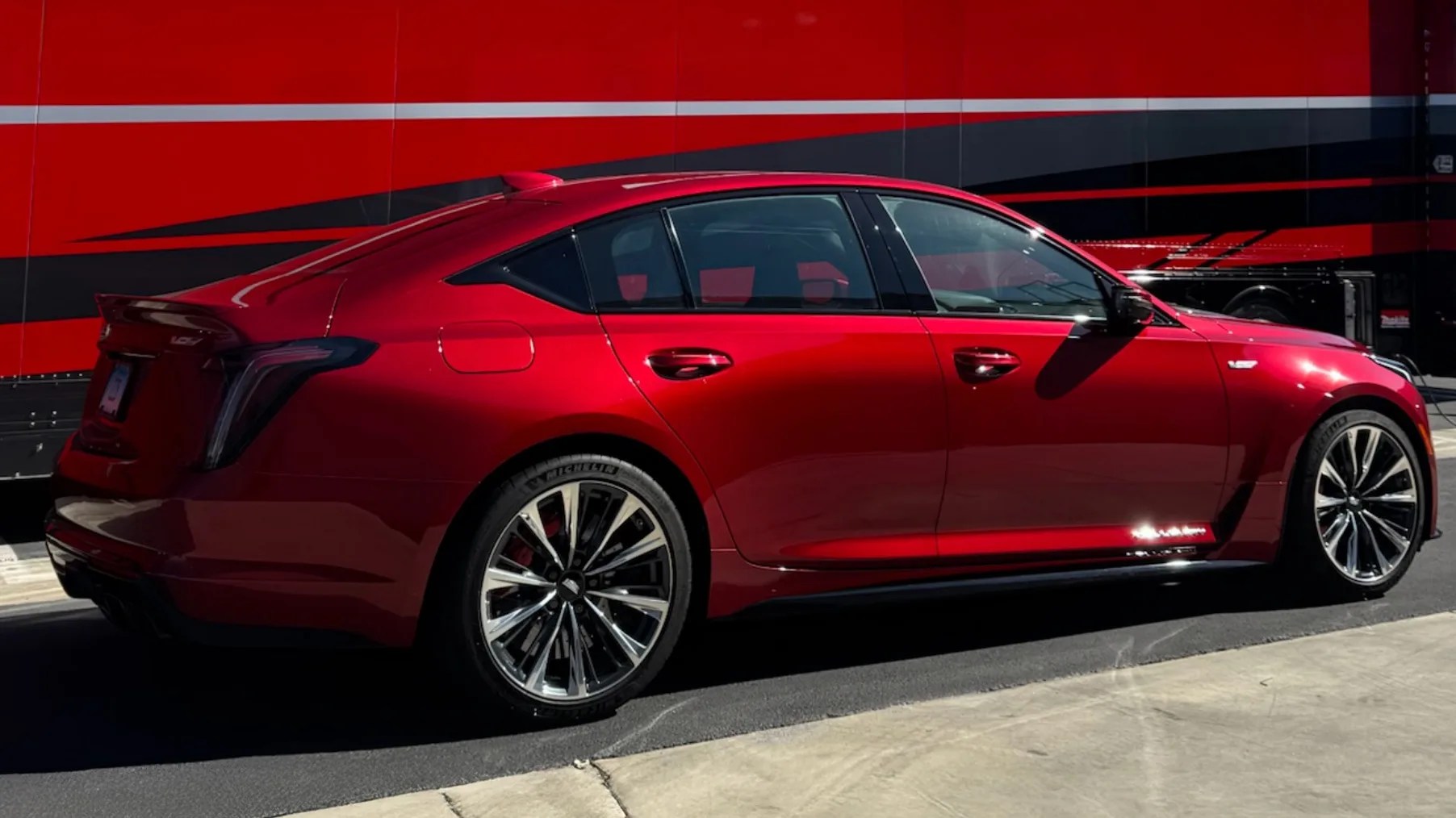 Red sporty sedan with black and silver alloy wheels parked on asphalt near a red and black trailer.