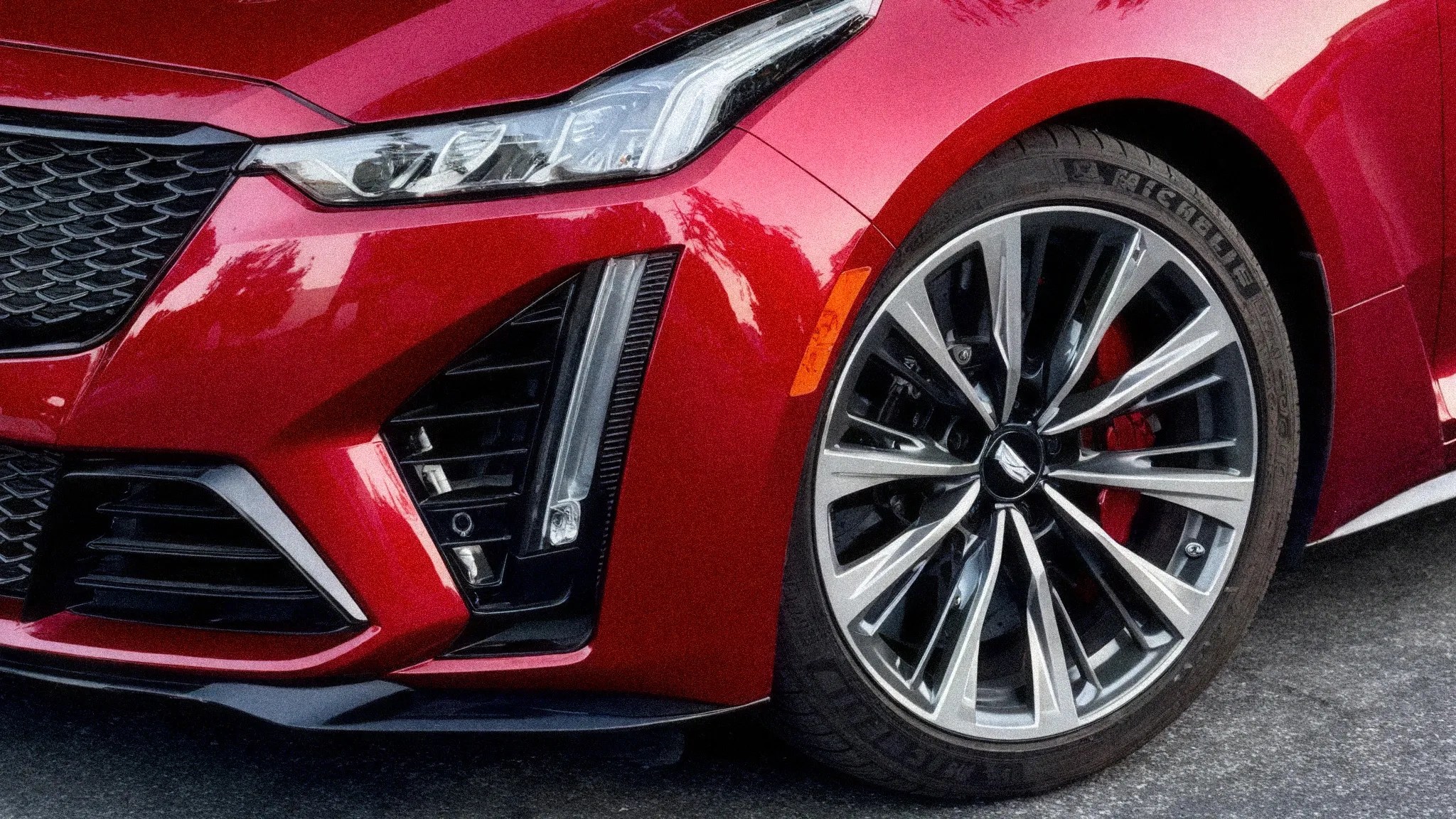 Close-up of a red car's front left headlight, grille, and multi-spoke alloy wheel with a low-profile tire.