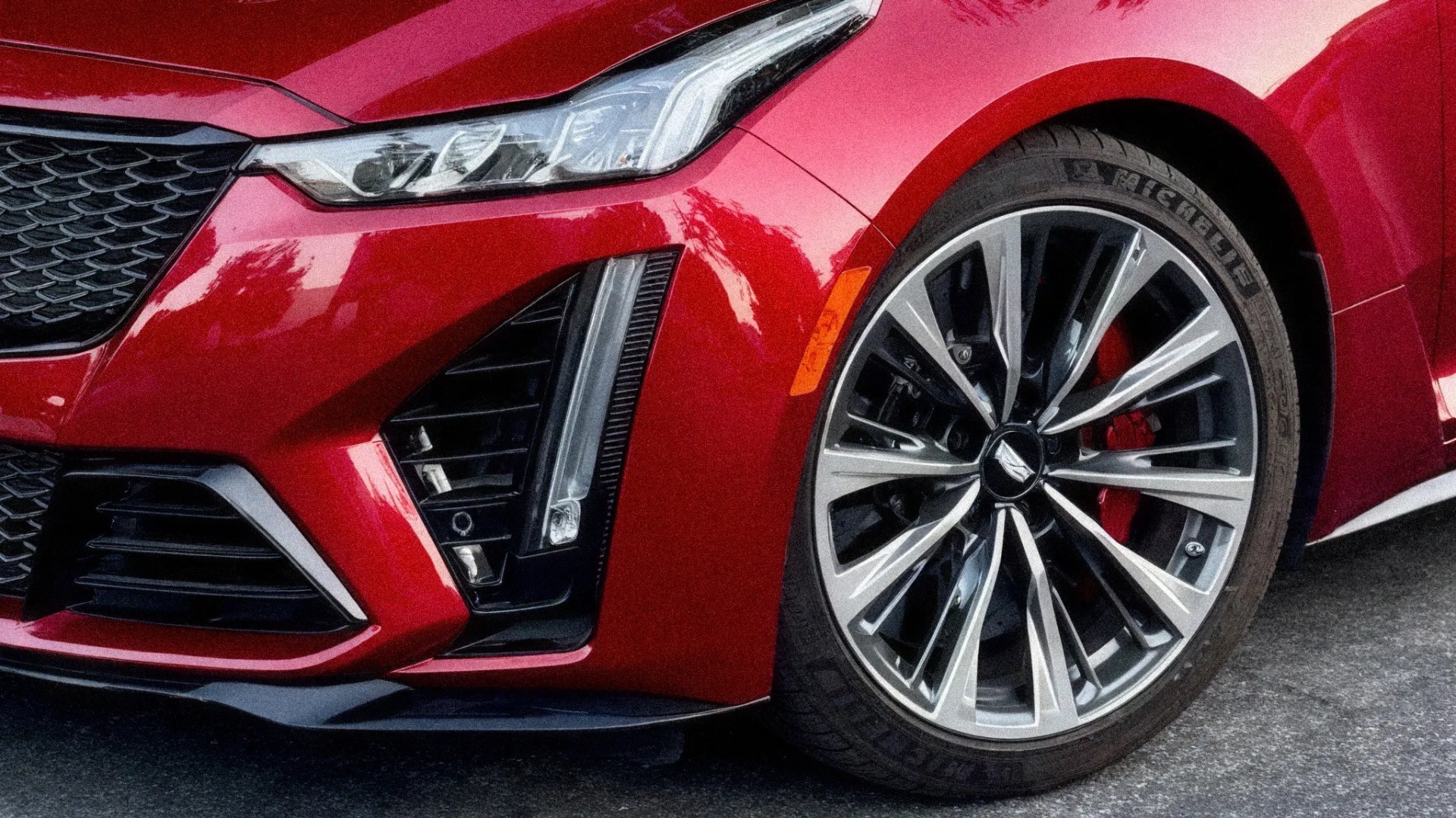 Close-up of a red car's front left headlight, grille, and multi-spoke alloy wheel with a low-profile tire.