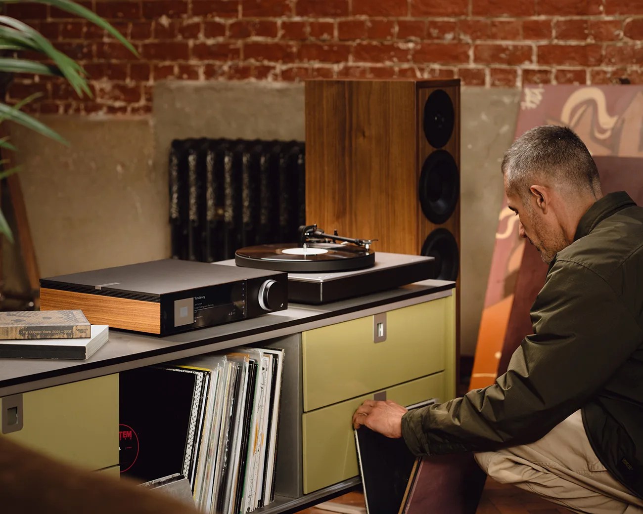 Man in a green jacket kneeling by a cabinet with vinyl records, next to a turntable and wooden speaker on top.