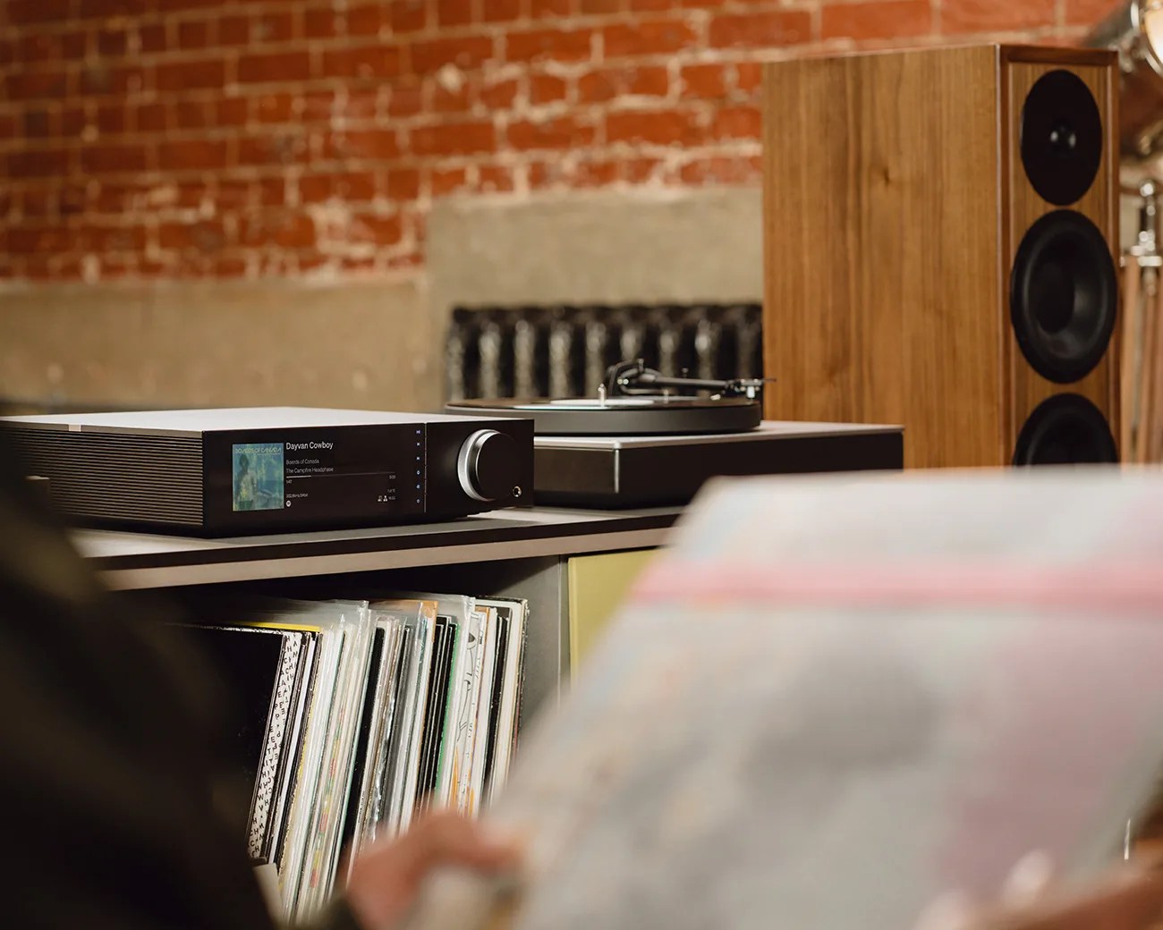 Black audio receiver with digital display and large knob on a shelf next to a wooden speaker and vinyl records.
