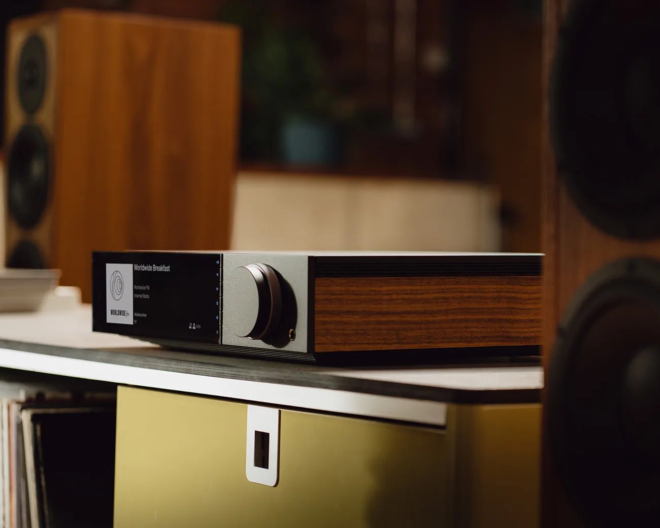 Audio receiver with wood panel sides and a digital display showing "Worldwide Breakfast" on a shelf between two wooden speakers.