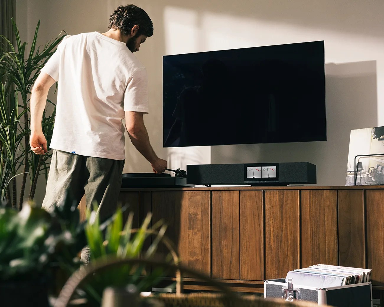 Man adjusting a record player on a wooden cabinet with a black soundbar and wall-mounted TV above.