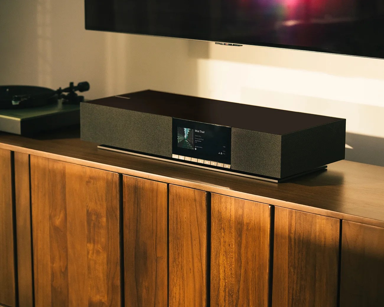 Rectangular black soundbar with fabric-covered speakers and a central digital display on a wooden cabinet.