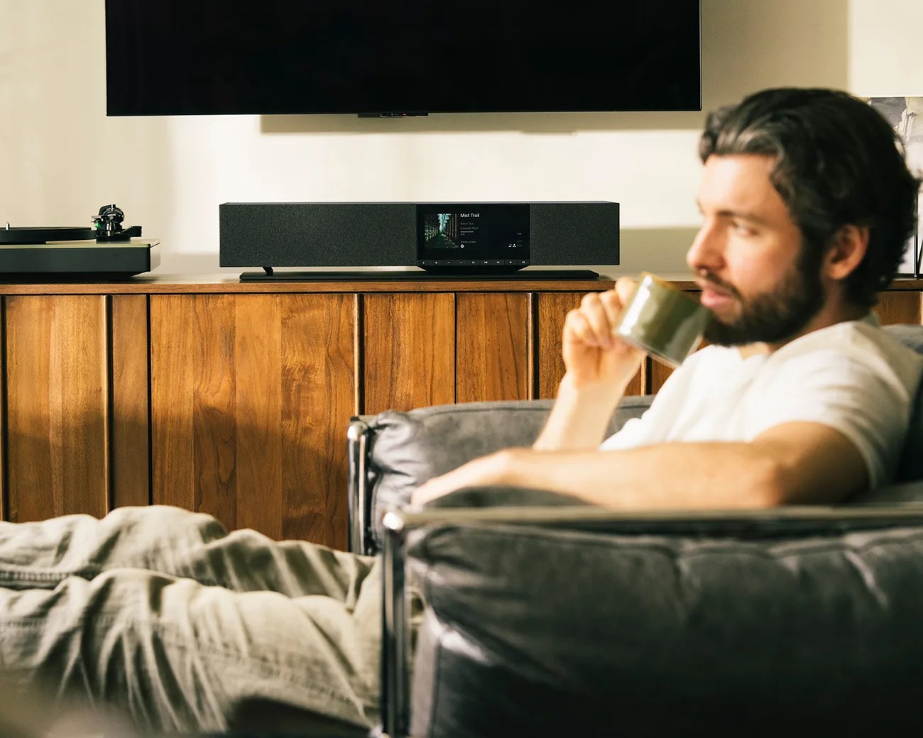 Man sitting on a black leather chair drinking from a green cup with a wooden cabinet and audio system in the background.