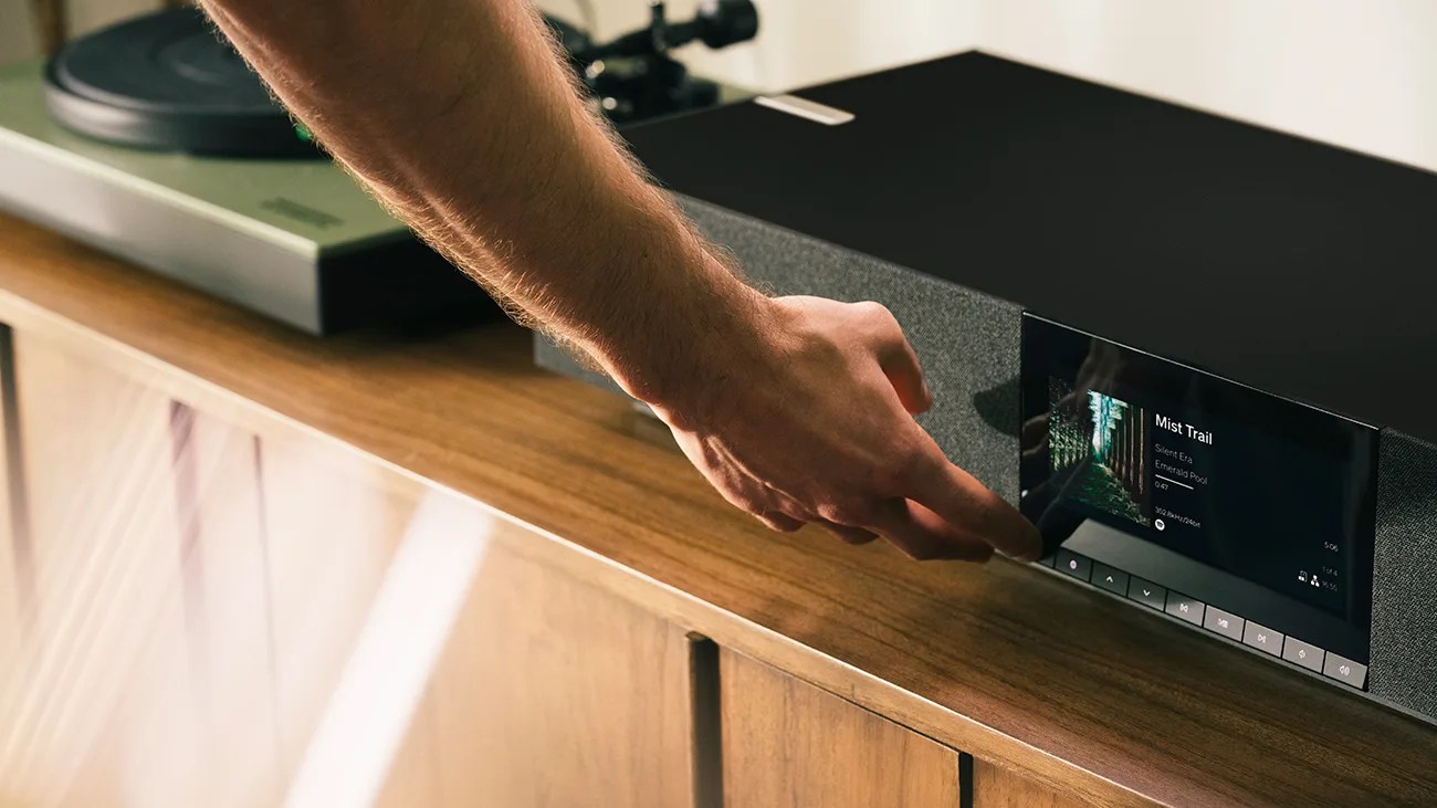 Hand adjusting controls on a black soundbar with a digital display on a wooden cabinet.