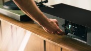 Hand adjusting controls on a black soundbar with a digital display on a wooden cabinet.