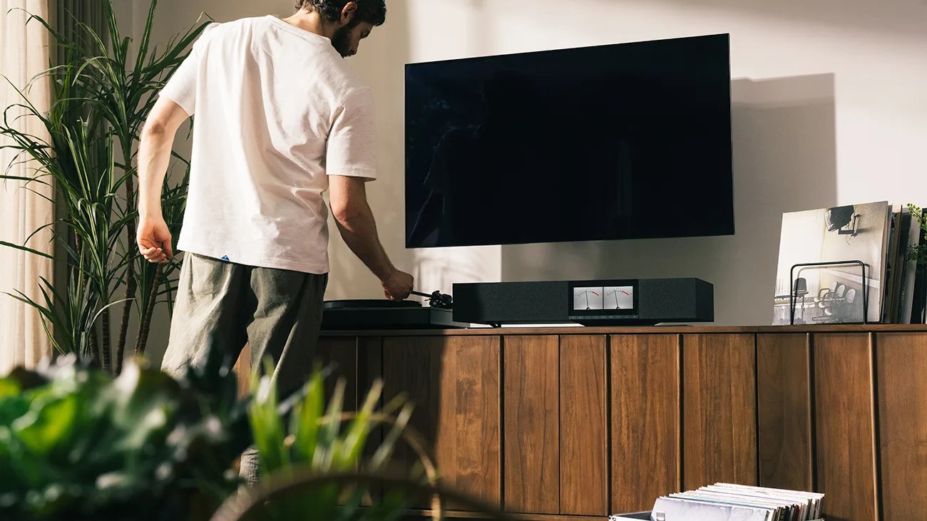 Man adjusting a black turntable on a wooden cabinet with a wall-mounted flat-screen TV above.