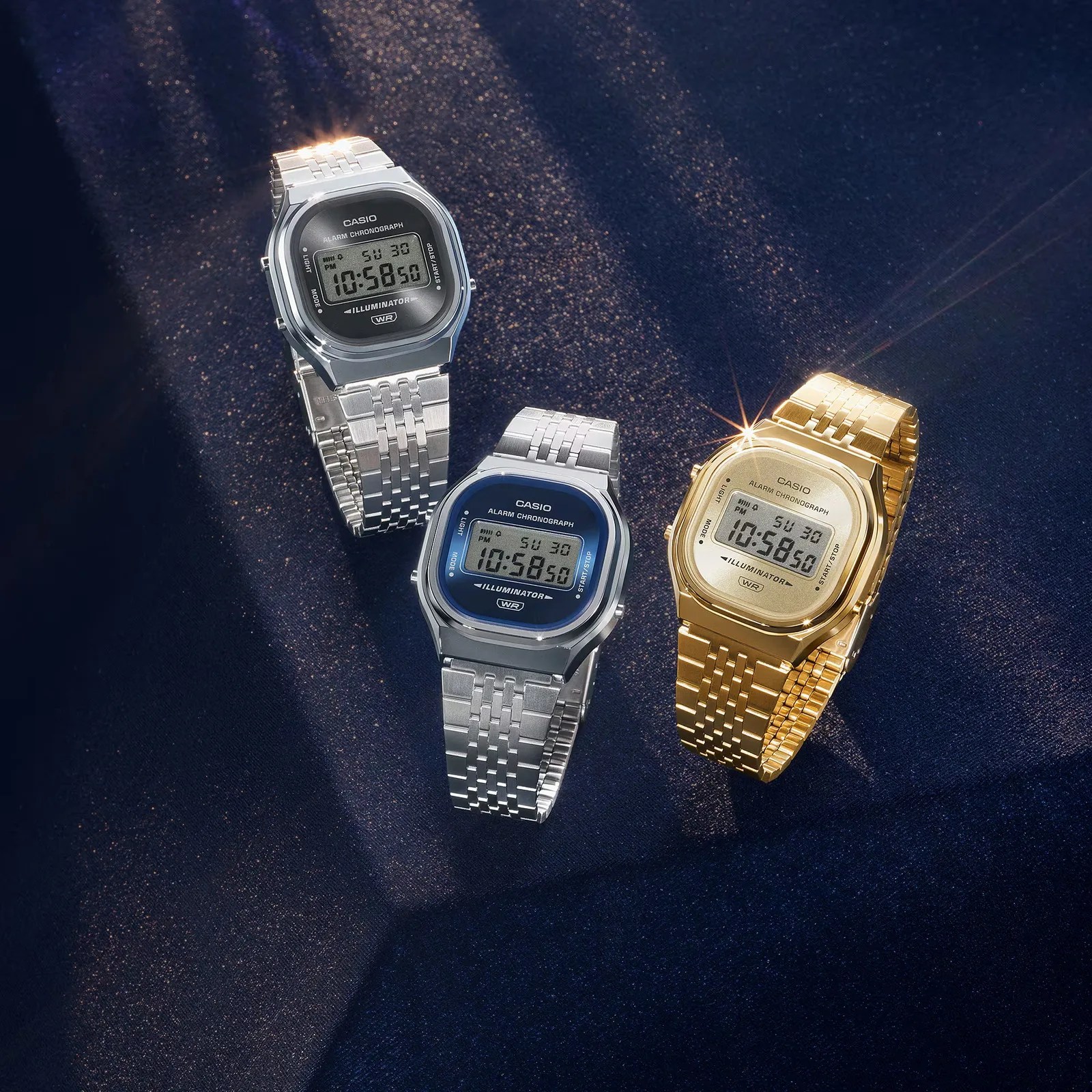Three Casio digital wristwatches with metal bands in silver, silver with blue face, and gold on dark fabric.