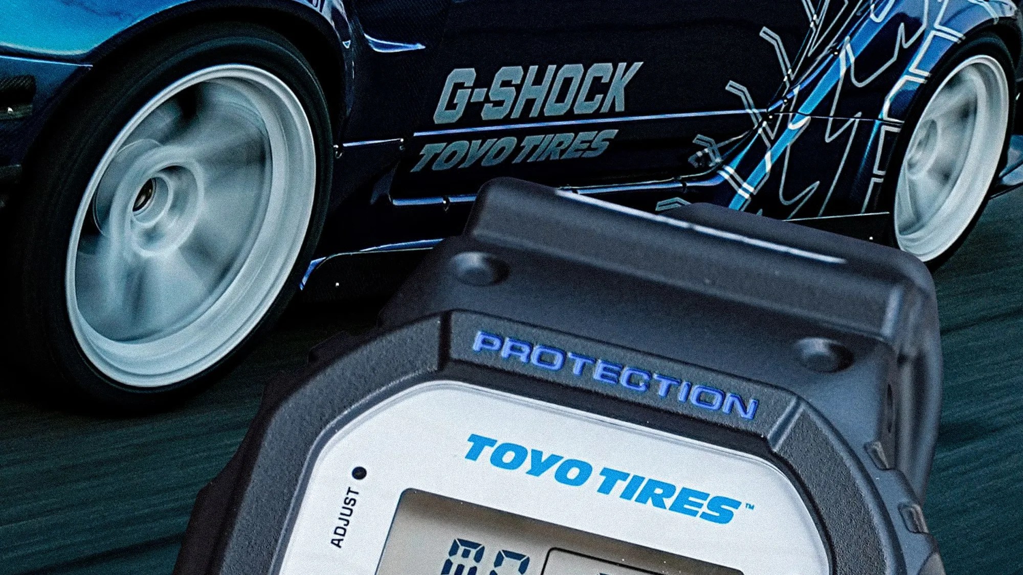 Close-up of a black G-Shock watch with "TOYO TIRES" branding in front of a blue car with spinning white wheels and "G-SHOCK TOYO TIRES" decals.